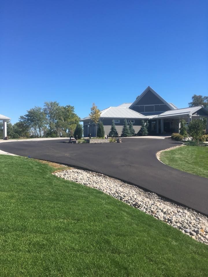 A gray, multi-gabled house with a large, freshly paved asphalt driveway and a decorative rock border under a blue sky.