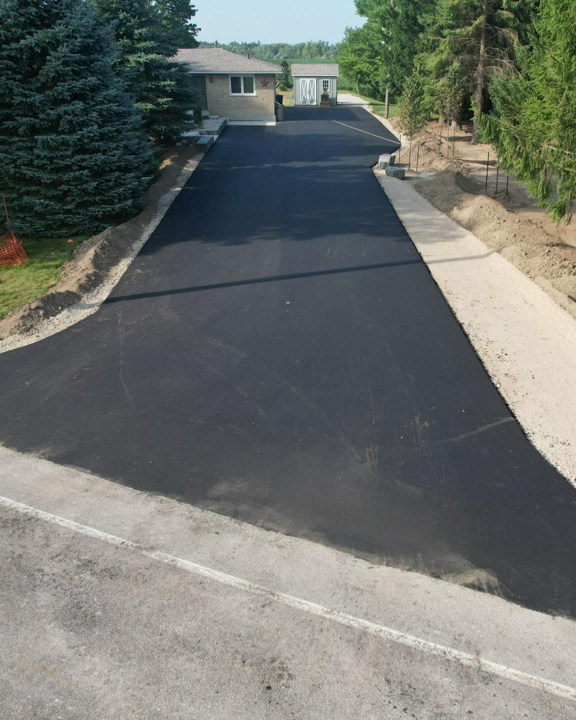 A newly paved black asphalt driveway leads up to a residential house, framed by trees and a concrete walkway.