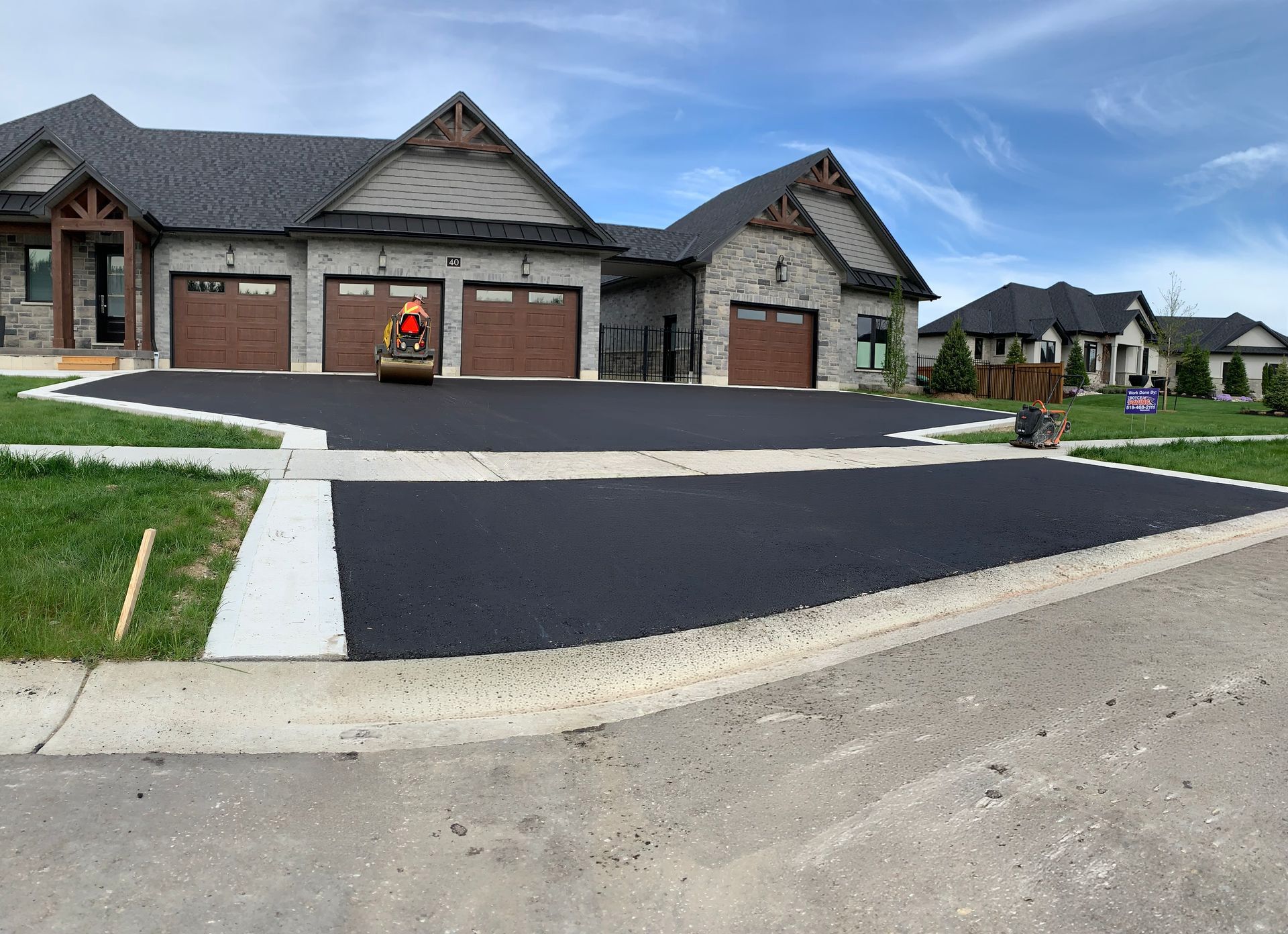 A freshly paved asphalt driveway in front of a stone residential house, with a construction roller parked near the garage.