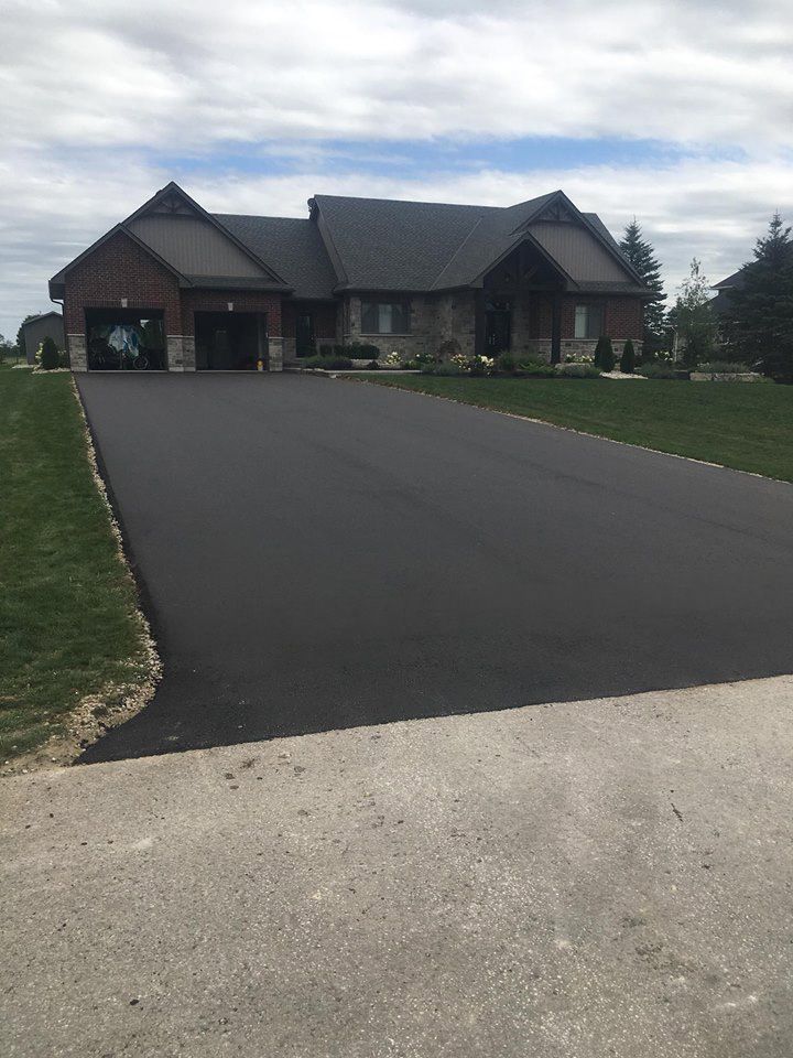 A newly paved dark asphalt driveway leads to a large suburban house with a multi-car garage on a sunny day.