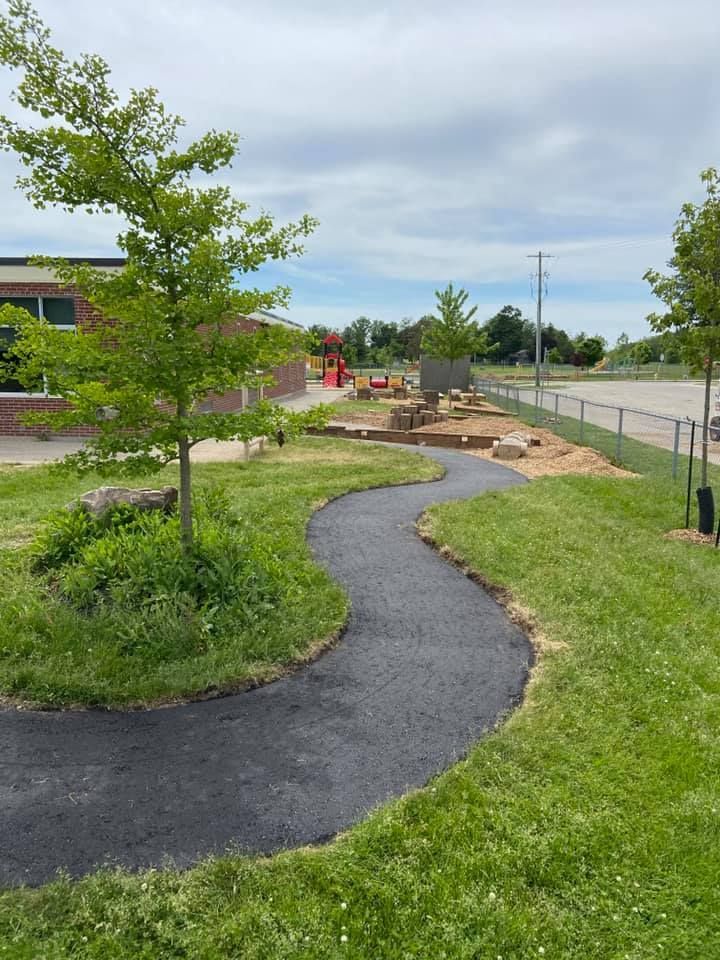 A curving paved walkway winds through a green grass yard with small trees and landscaping rocks near a school building.