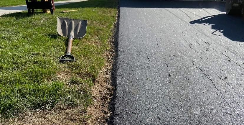 A shovel rests on the grass beside a freshly paved asphalt driveway edge.