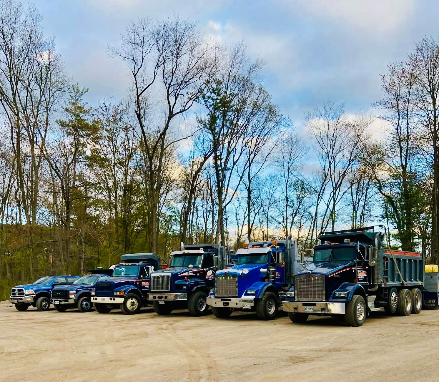 A line of blue dump trucks and a pickup truck parked on a gravel lot in front of trees under a cloudy sky.