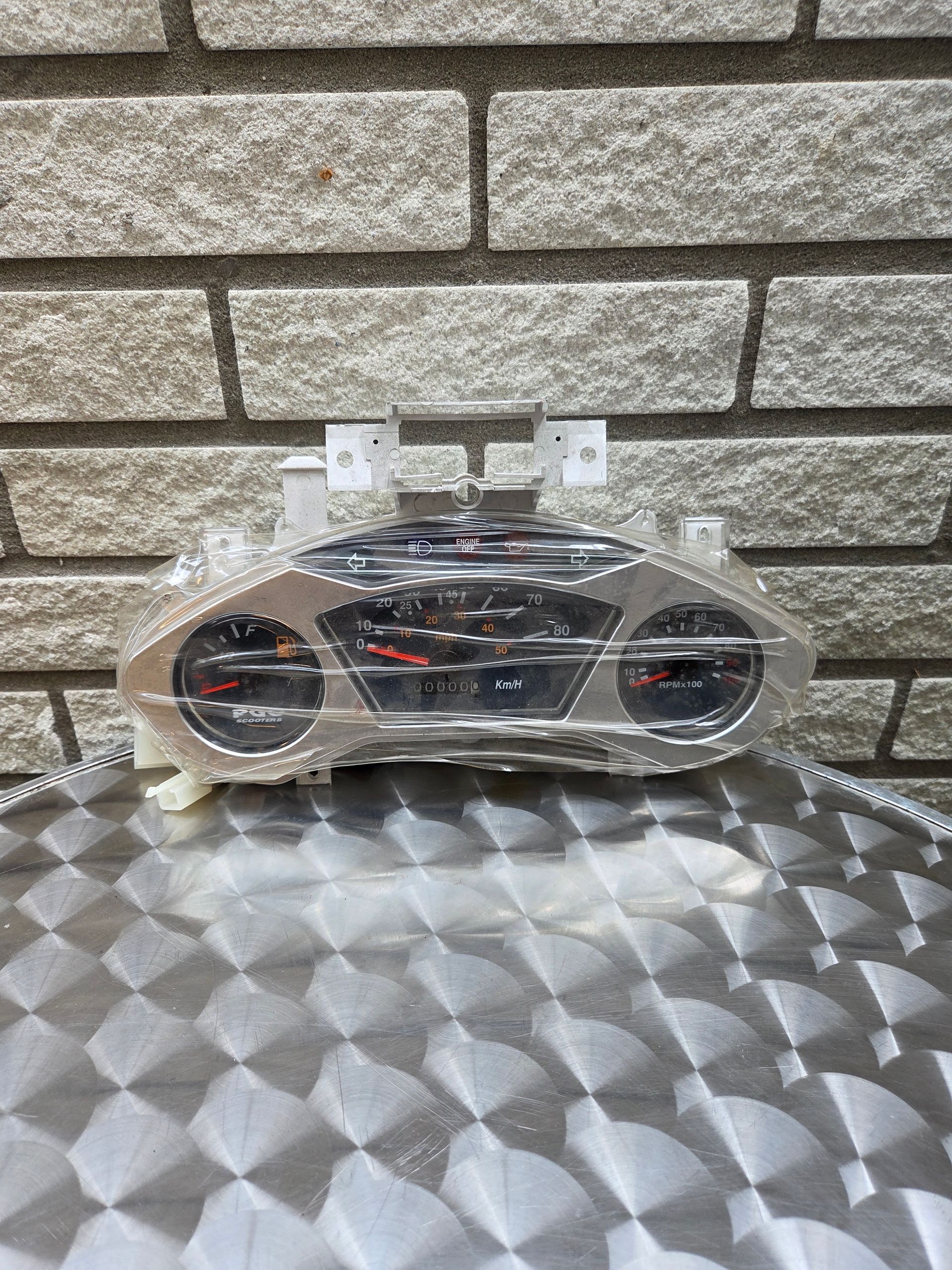 Een met transparant plastic bekleed instrumentenpaneel op een zilverkleurige ondergrond met patroon, tegen een bakstenen muur.