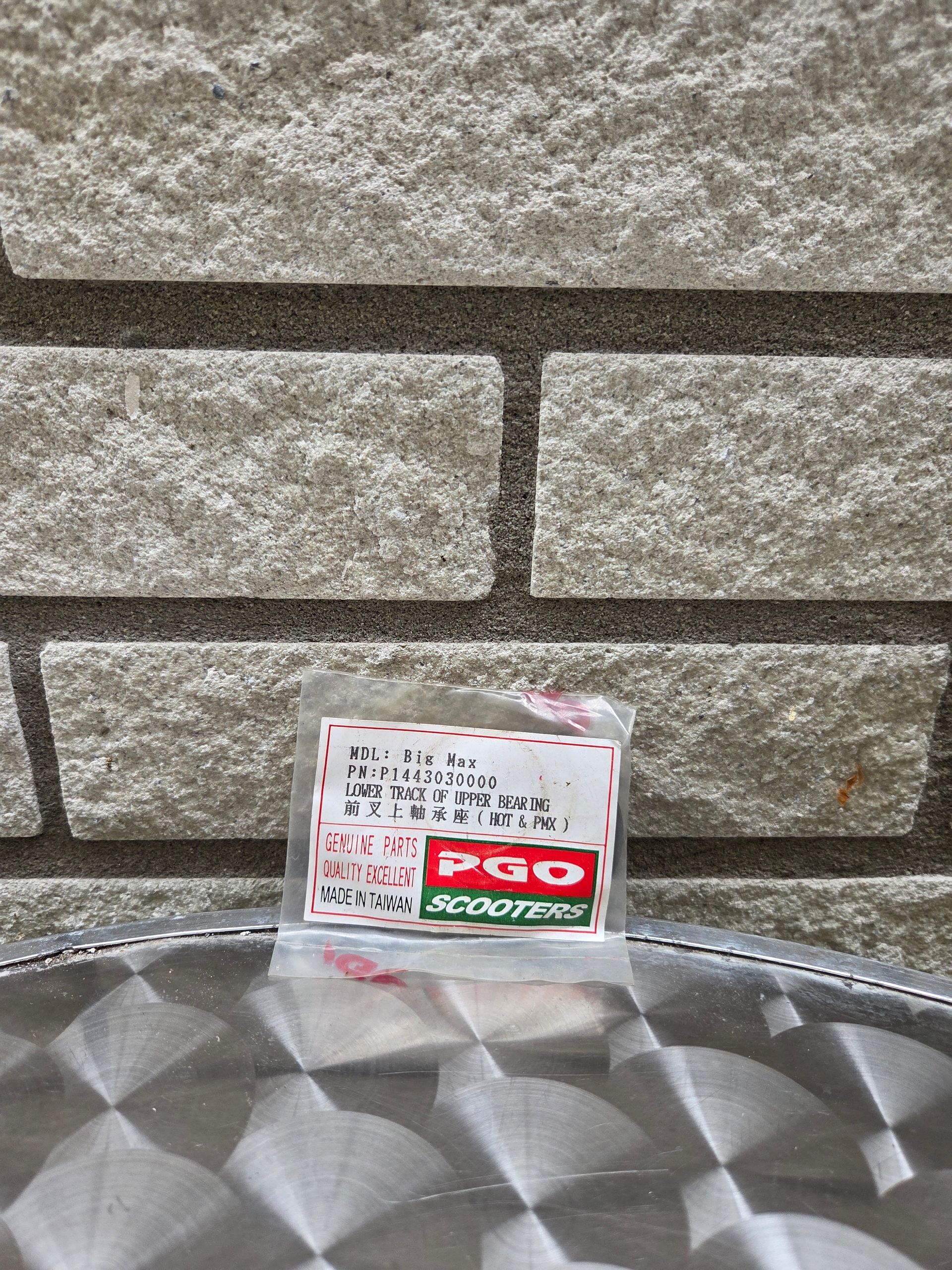Klein bordje met Japanse tekst en een rood logo, tegen een stenen muur geplaatst boven een zilverkleurig, gestructureerd oppervlak.