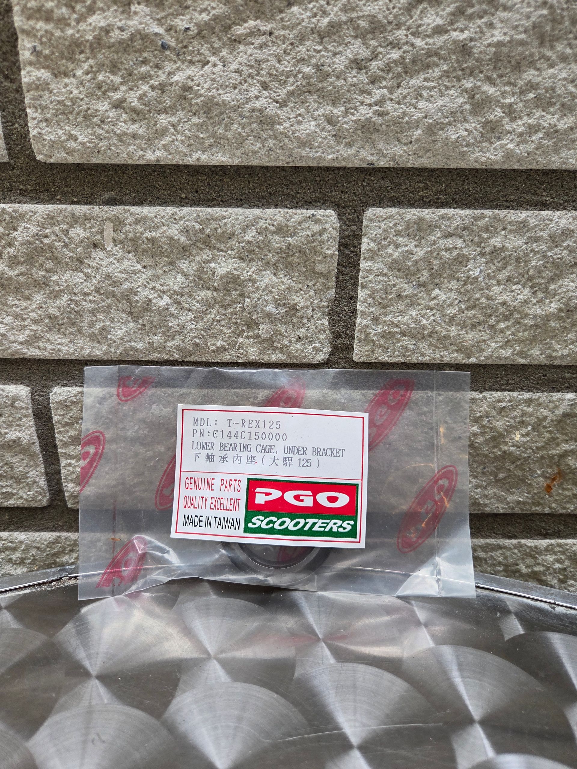Een kleine PGO Scooters-doos op een metalen ondergrond tegen een stenen muur.