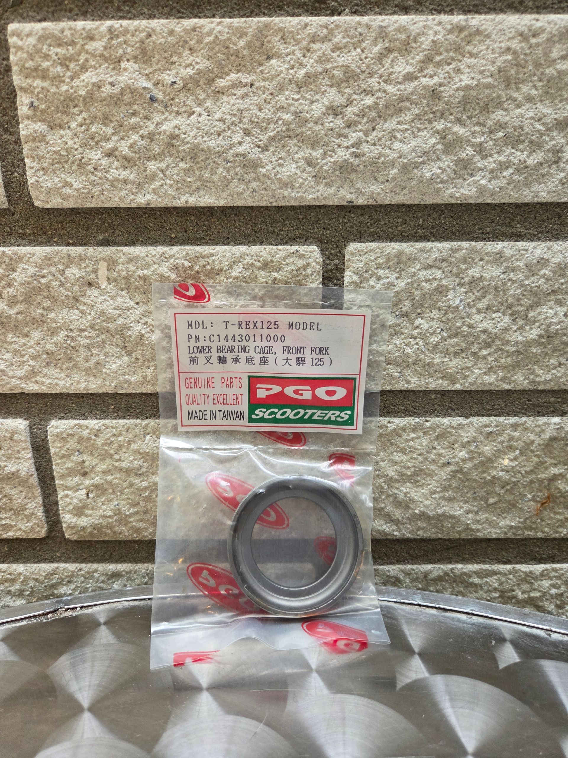 Een kleine, verzegelde metalen ring in een plastic verpakking, die op een reflecterend metalen oppervlak tegen een bakstenen muur ligt.