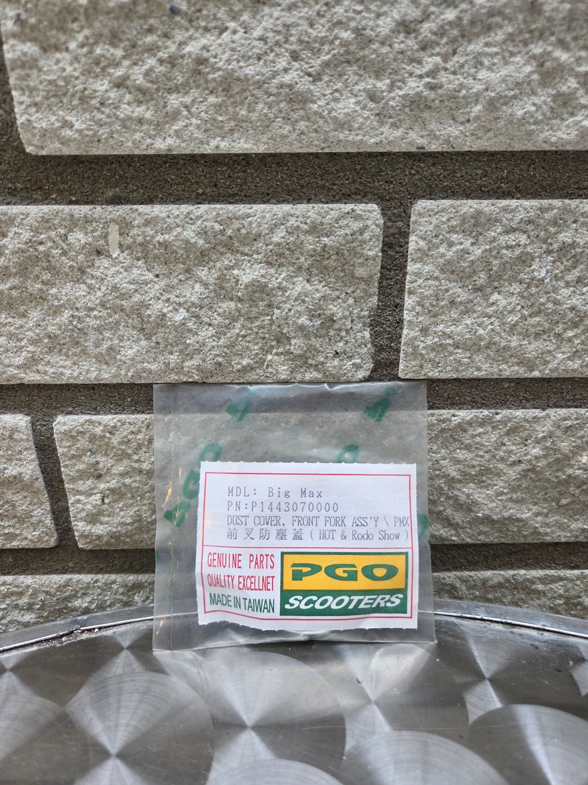 Klein, doorzichtig tasje met een sticker en het PGO Scooters-logo, leunend tegen een stenen muur op een metalen ondergrond.