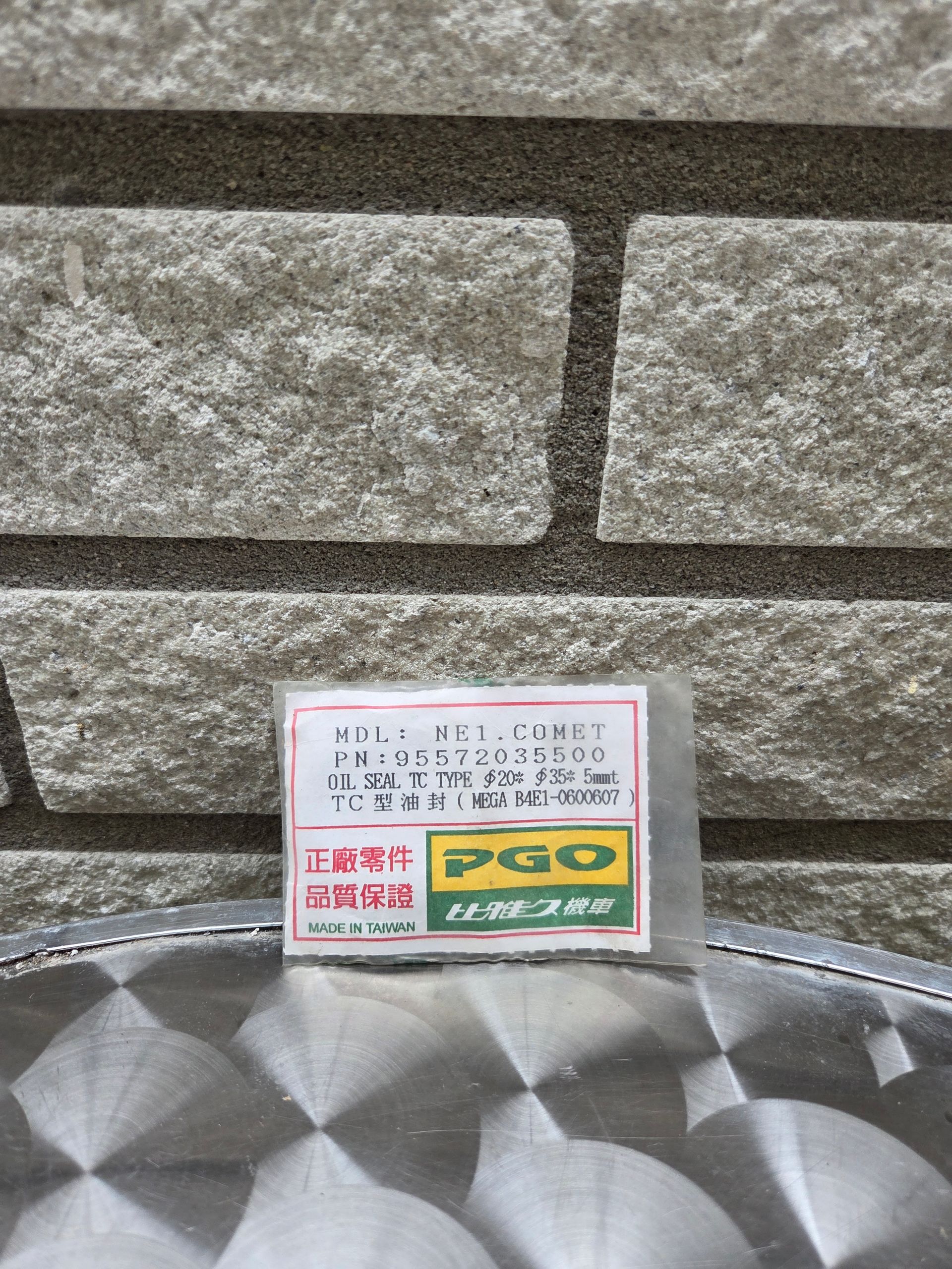 Klein bordje op een stenen muur boven een reflecterend metalen oppervlak, met Japanse tekst en een groen-geel logo.