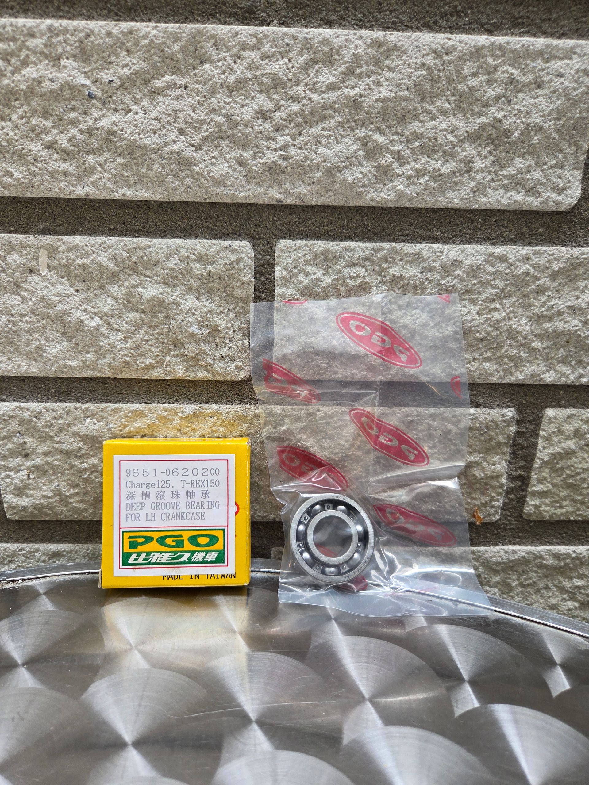 Klein geel doosje en een afgesloten plastic zak met een metalen lager op een reflecterend oppervlak tegen een bakstenen muur.