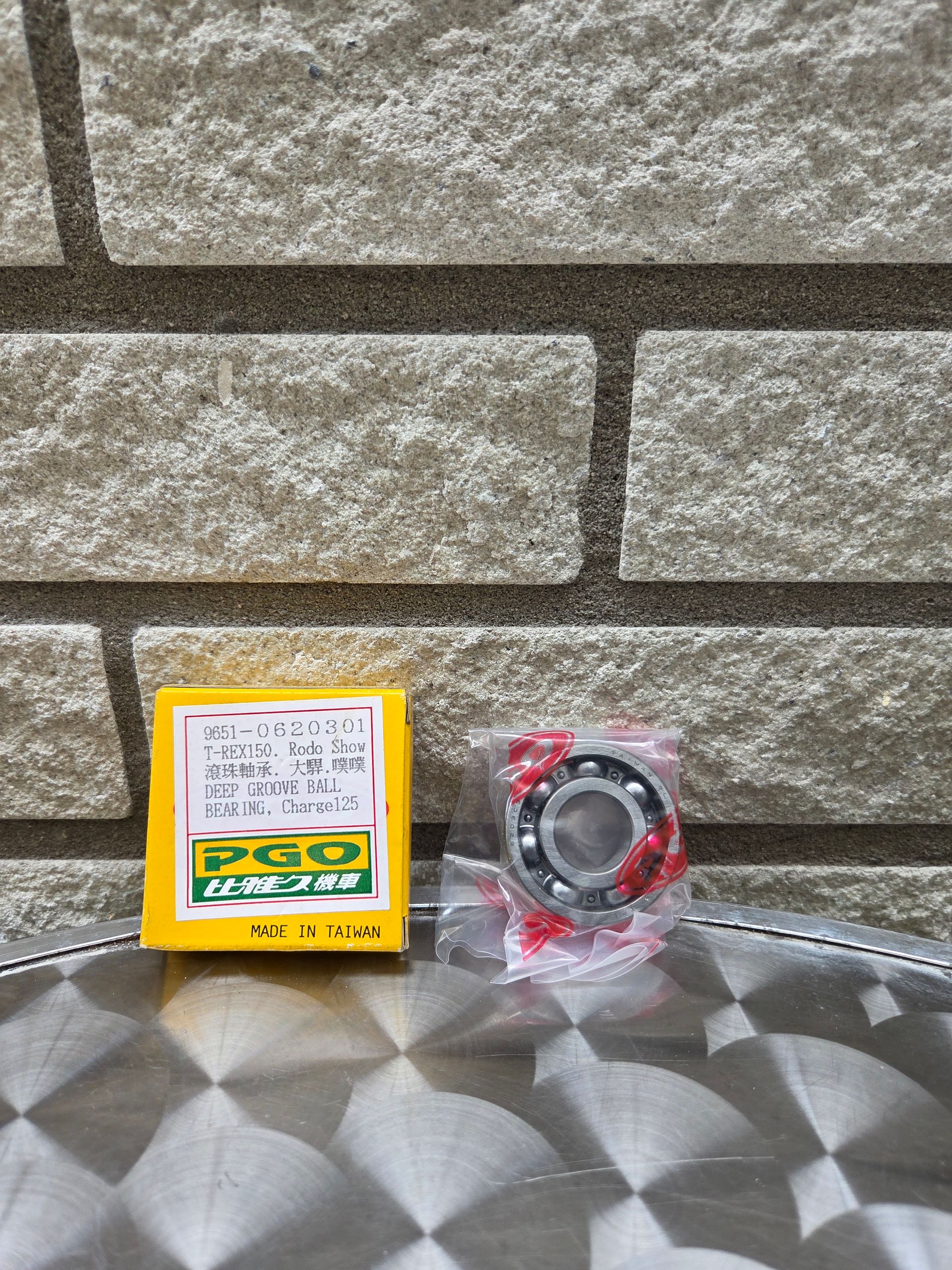 Een gele onderdelendoos en een in plastic verpakt lager op een grijs, bewerkt oppervlak tegen een stenen muur.