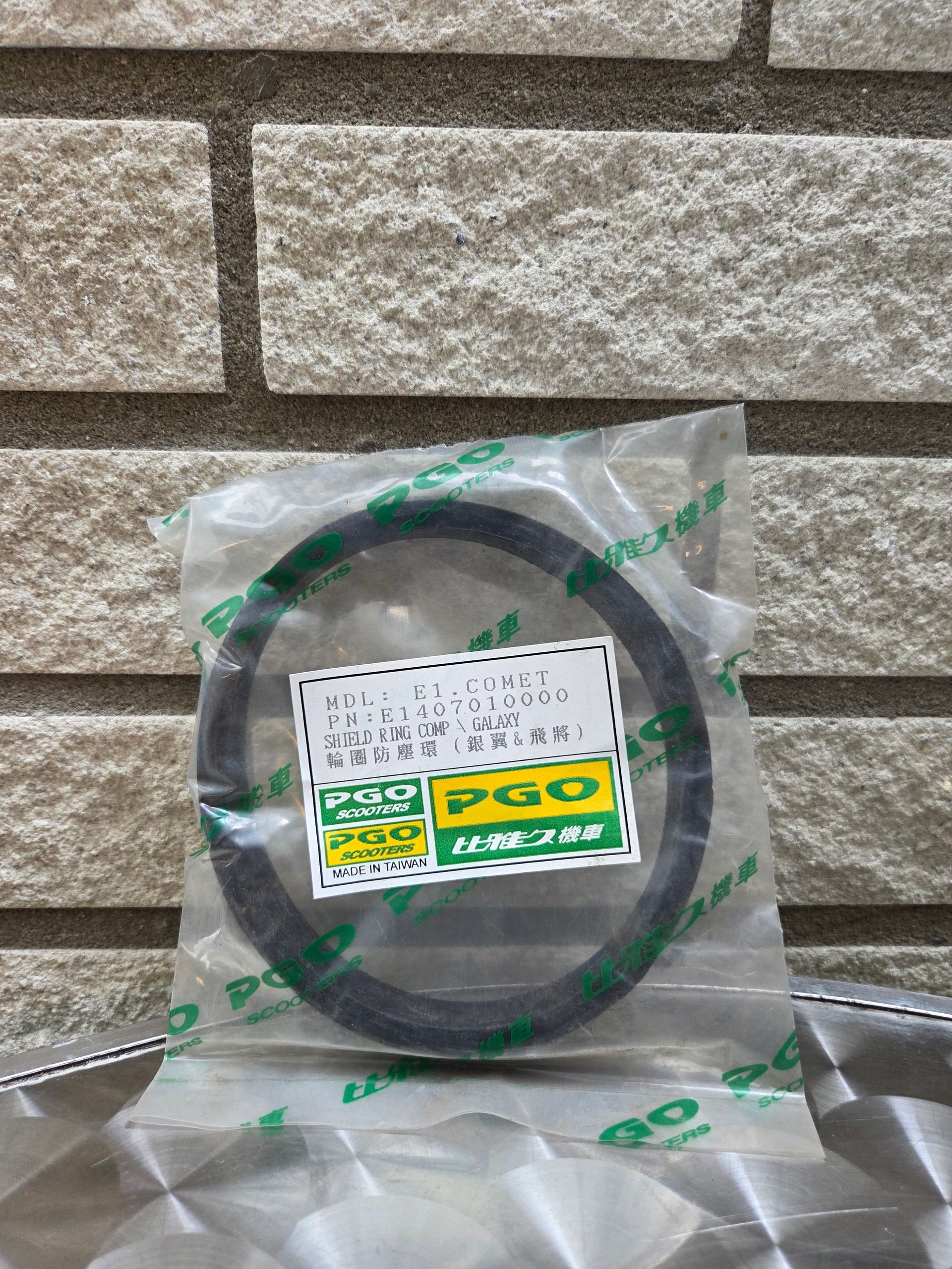 Doorzichtige plastic zak met daarin een zwart ringvormig onderdeel met een wit en groen etiket, tegen een achtergrond van een stenen muur.