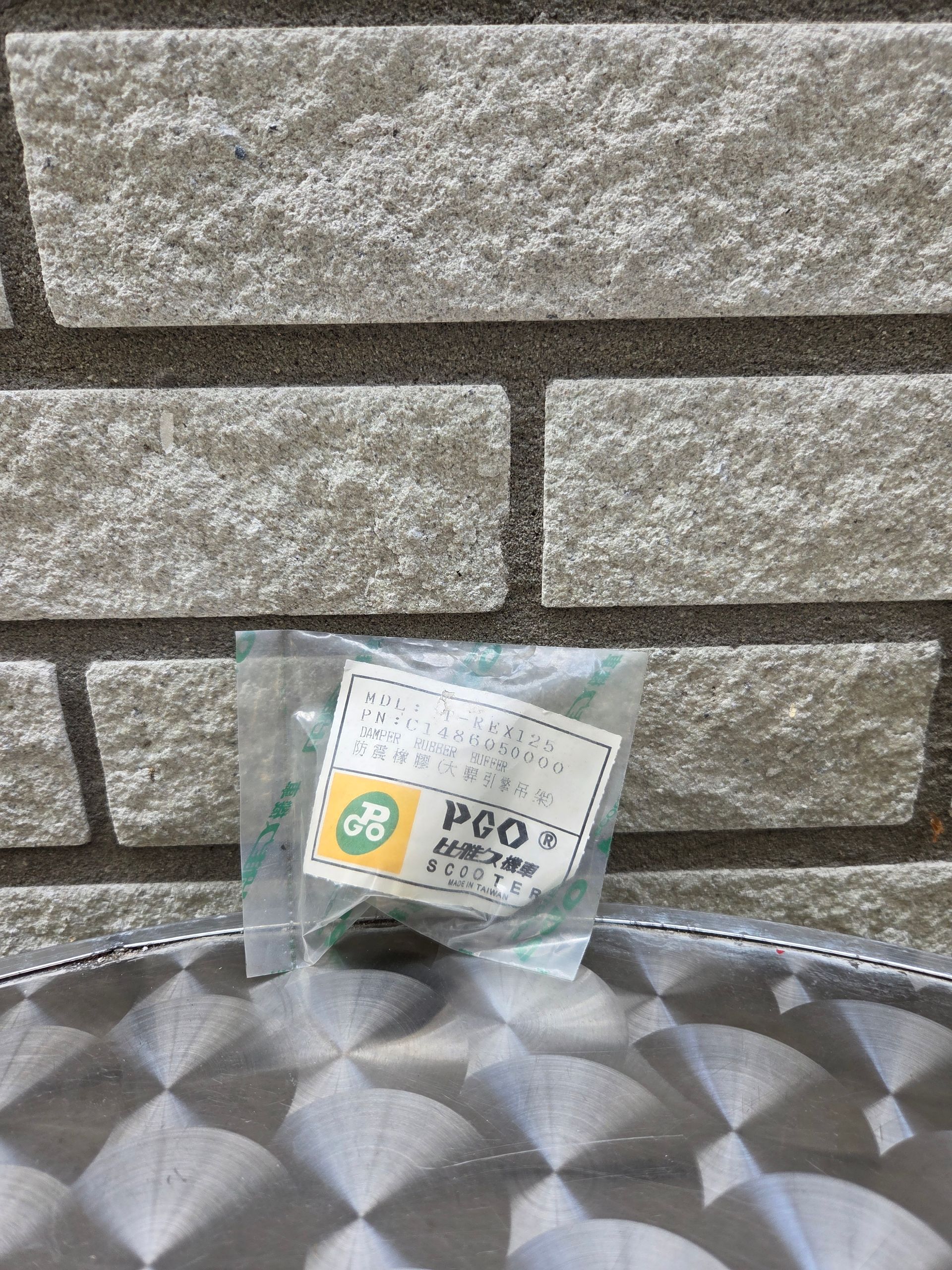 Klein, doorzichtig plastic zakje met een wit etiket en een geelgroen logo op een metalen ondergrond met patroon, tegen een stenen muur.