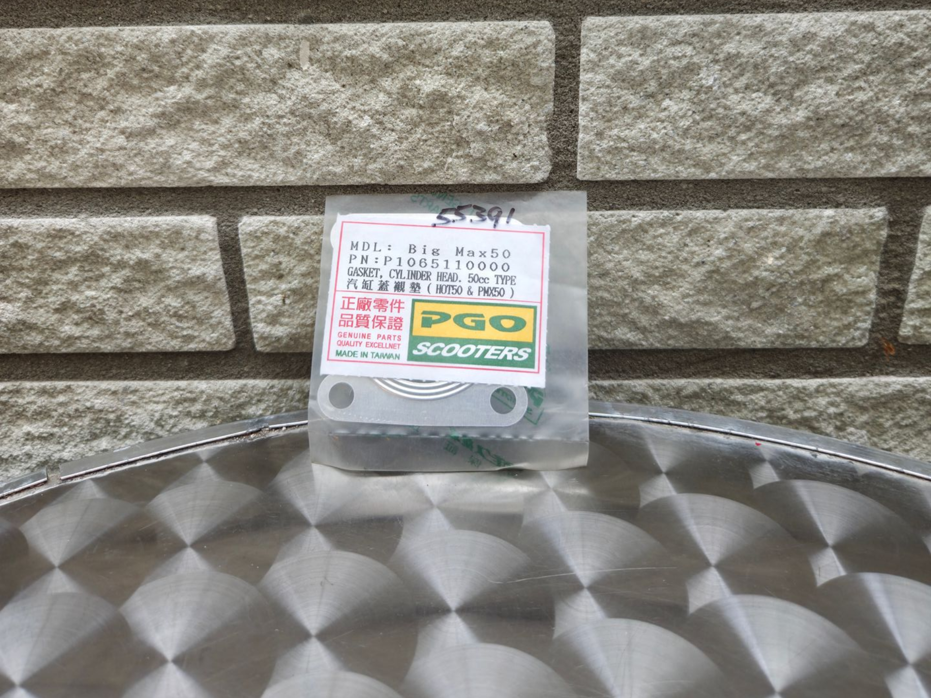 Een doorzichtige plastic doos met een kleurrijk etiket, die op een zilverkleurige, gestructureerde ondergrond tegen een bakstenen muur staat.