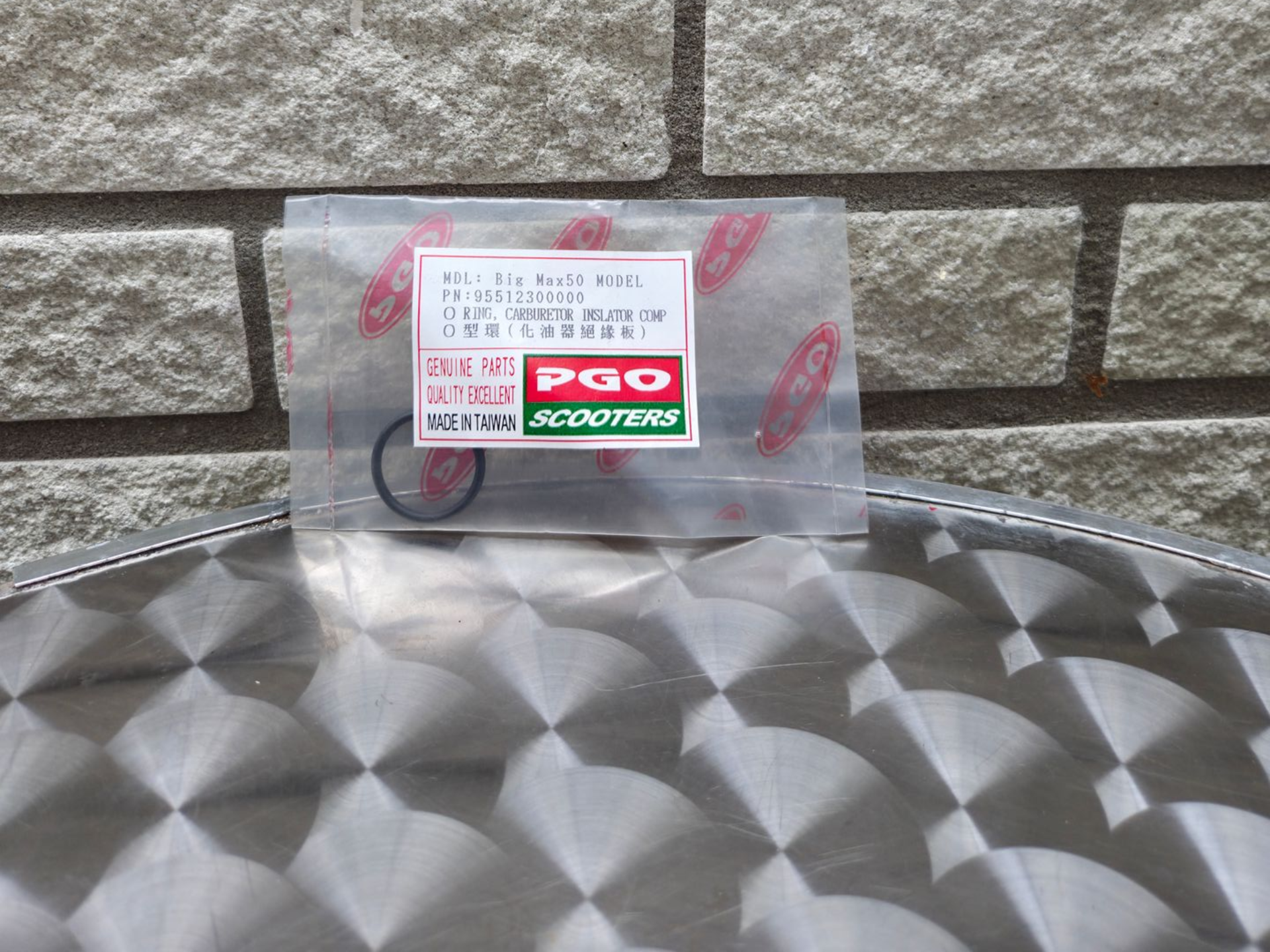 Een klein zwart-rood PGO-motoronderdeel in een doorzichtige plastic zak op een metalen tafel tegen een stenen muur.