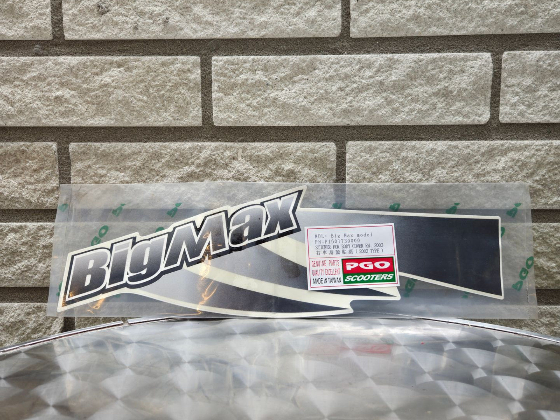 Een sticker met het BluMax-logo en een klein rood-groen etiket op een transparant plastic vel tegen een bakstenen muur.