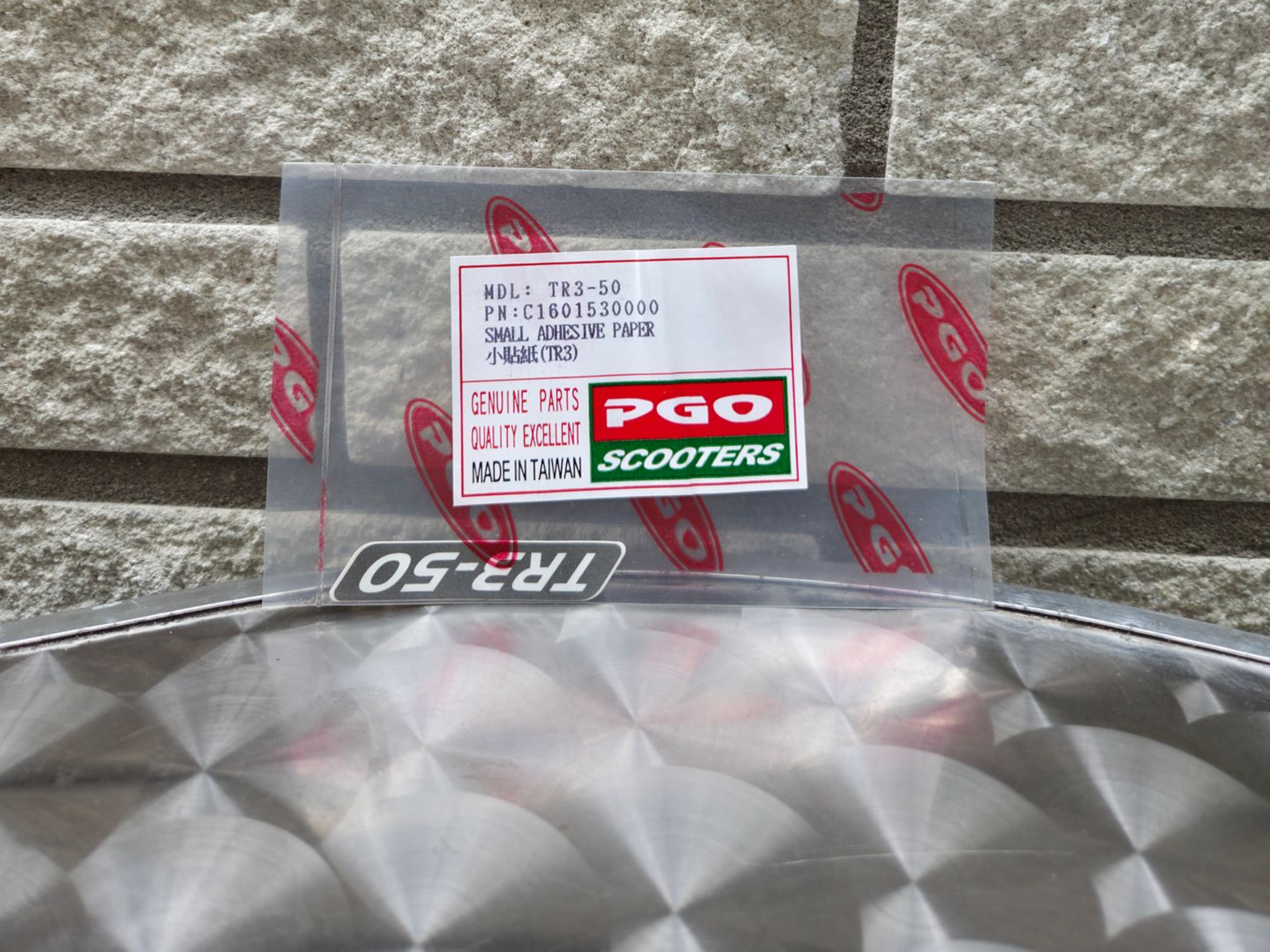 Doorzichtig plastic zakje met een PGO Scooters-label en rode logo's met een patroon op een zilverkleurig, gestructureerd oppervlak.