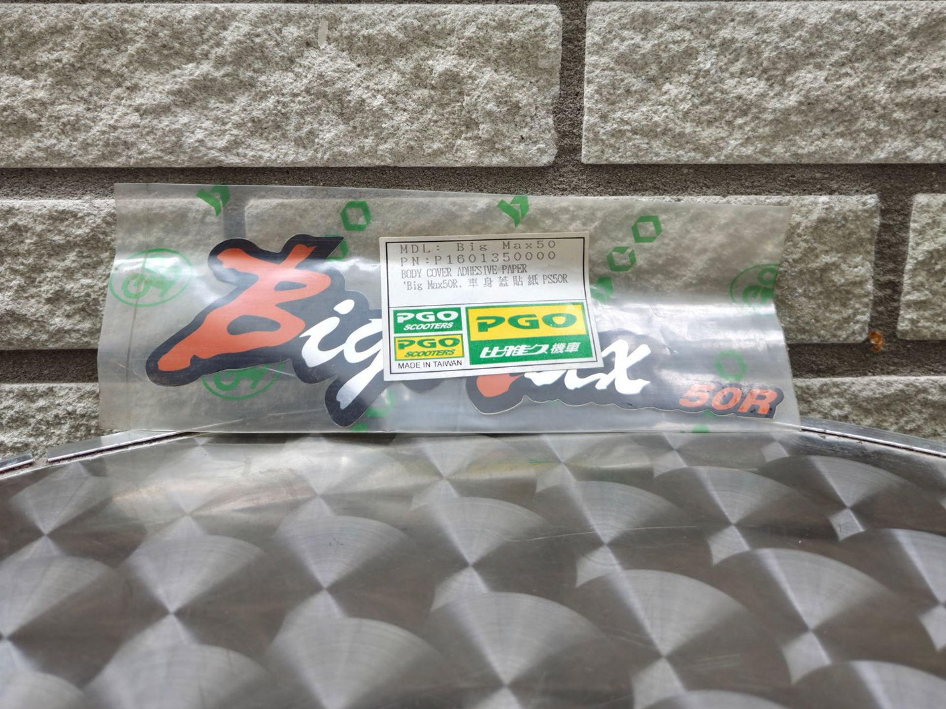 Een doorzichtig plastic zakje met een groen etiket ligt op een grijs, patroonrijk motorkap van een auto, voor een bakstenen muur.