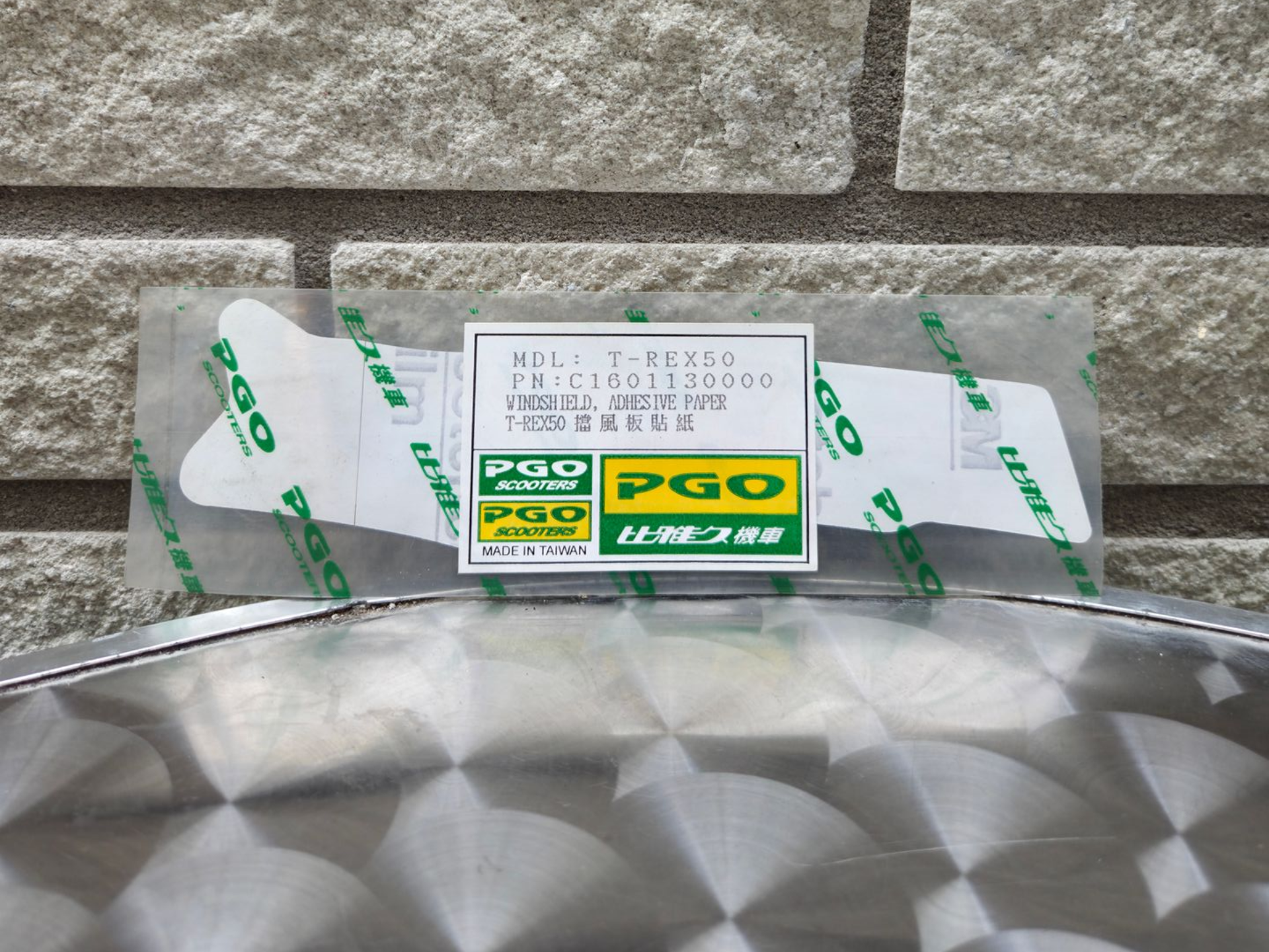 Een wit, in een doos verpakt artikel met groene PGO-etiketten, dat op een metalen ondergrond tegen een stenen muur staat.