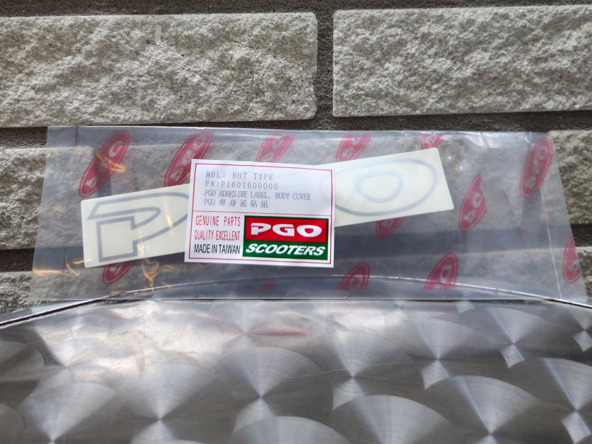 Doorzichtige plastic zak met scooterstickers en een PGO Scooters-label op een stenen ondergrond.