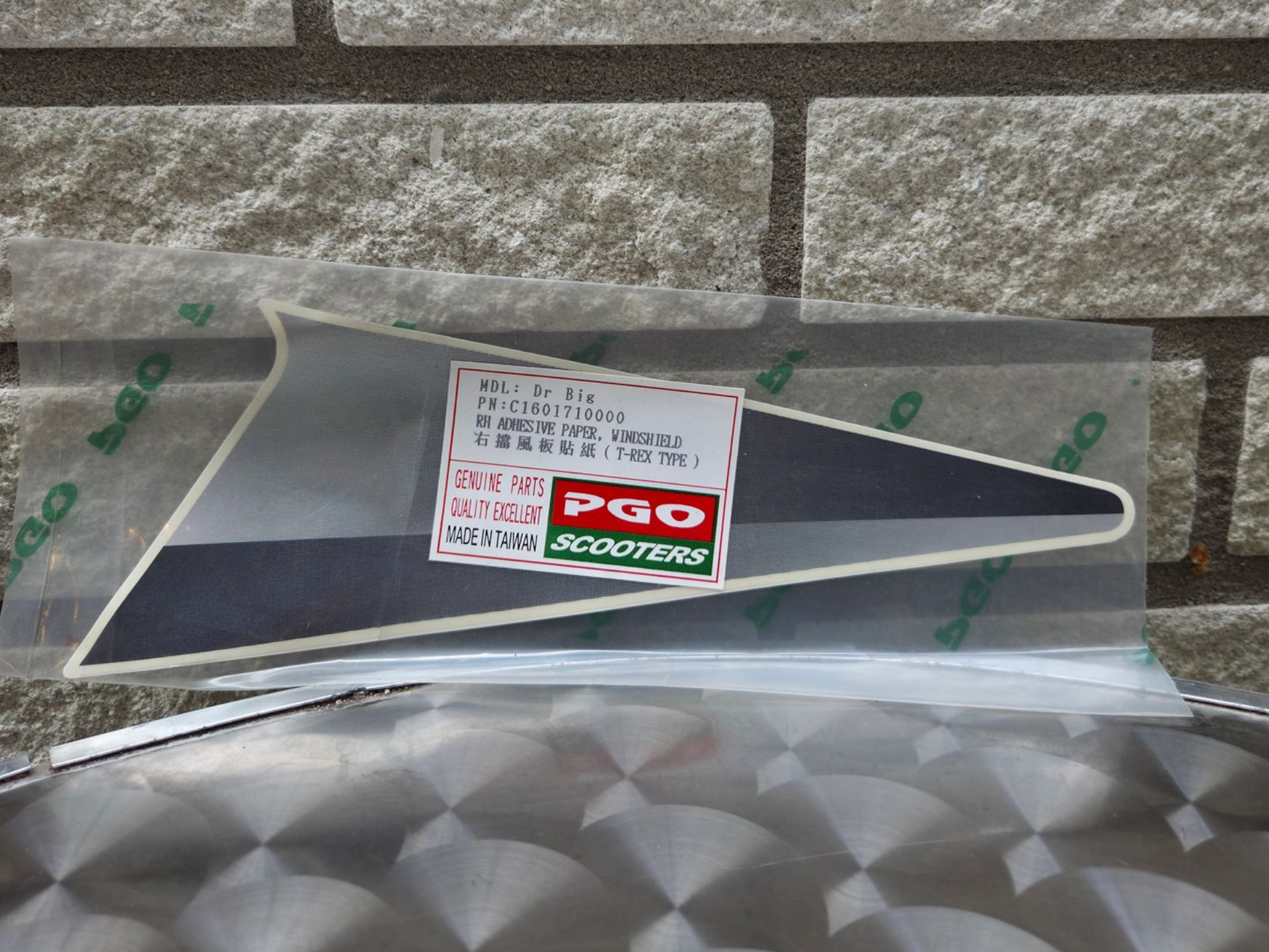 Rode driehoekige waarschuwingssticker met PGO-logo op een in plastic verpakte zilverkleurige ondergrond tegen een stenen muur.