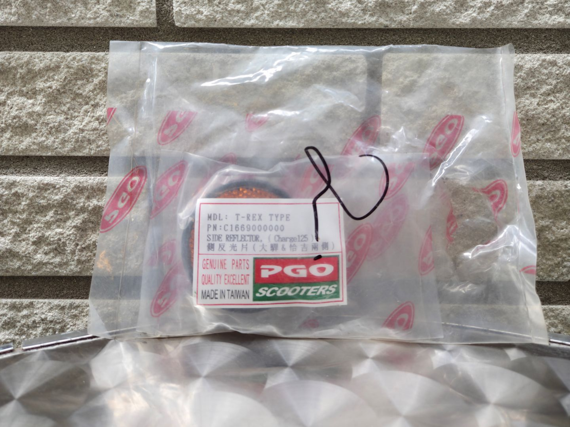 Doorzichtige plastic zak met een PGO Scooters-label en een zwarte draad op een stenen ondergrond.