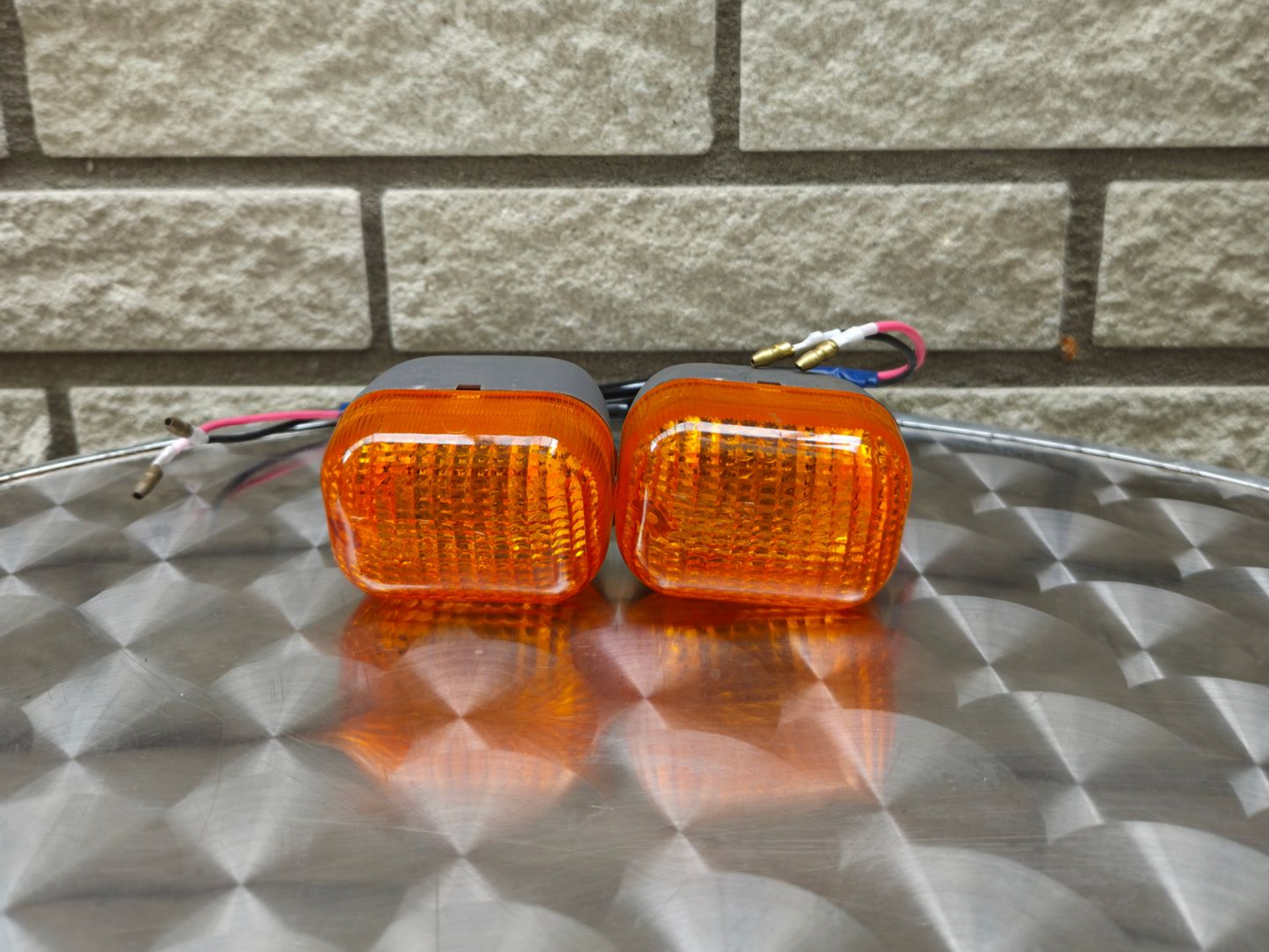 Twee amberkleurige, rechthoekige lampen met draden, die op een metalen tafel tegen een bakstenen muur staan.