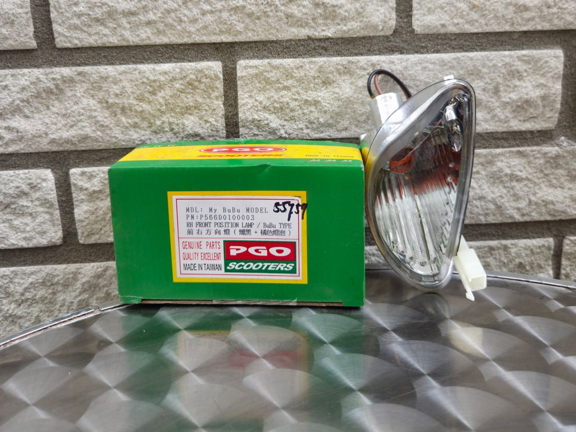 Groene PGO-elektrische scooterdoos naast een chromen koplamp op een metalen tafel tegen een bakstenen muur.