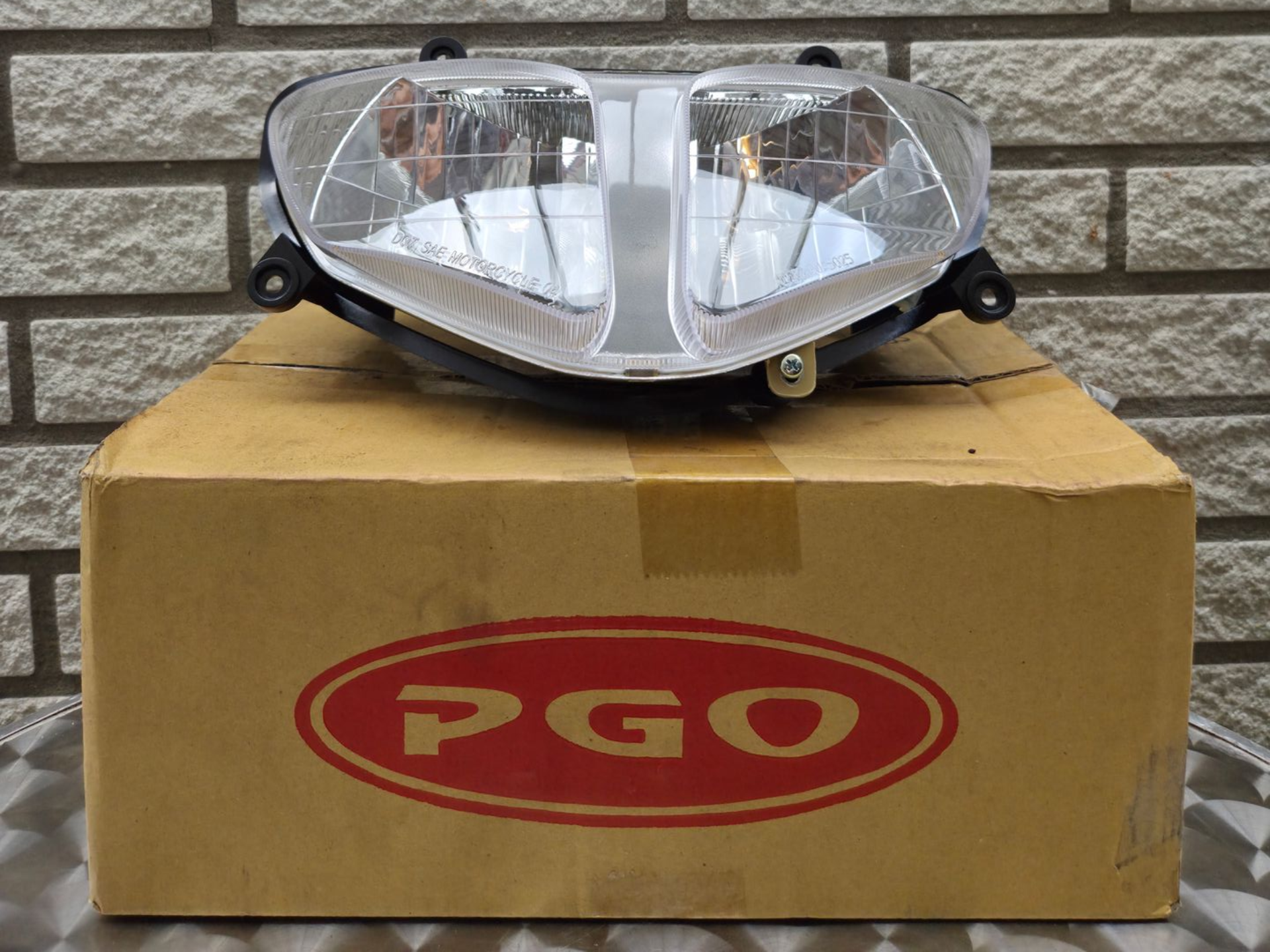 Motorfietskoplampunit op een PGO-kartonnen doos tegen een bakstenen muur.