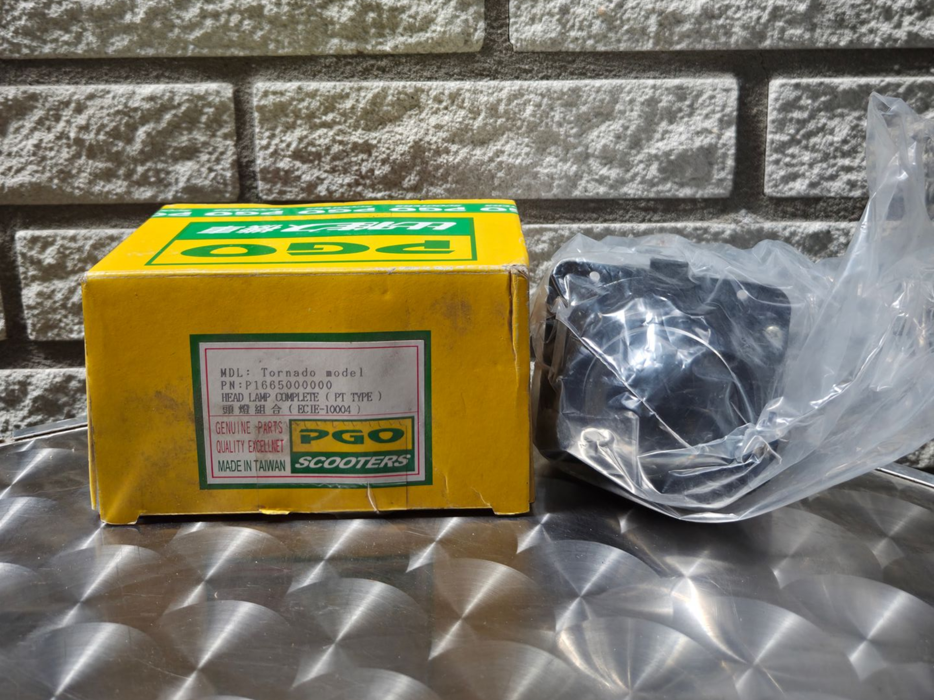 Gele doos met Japans etiket naast een zwart, rond onderdeel, verpakt in plastic, op een metalen oppervlak.