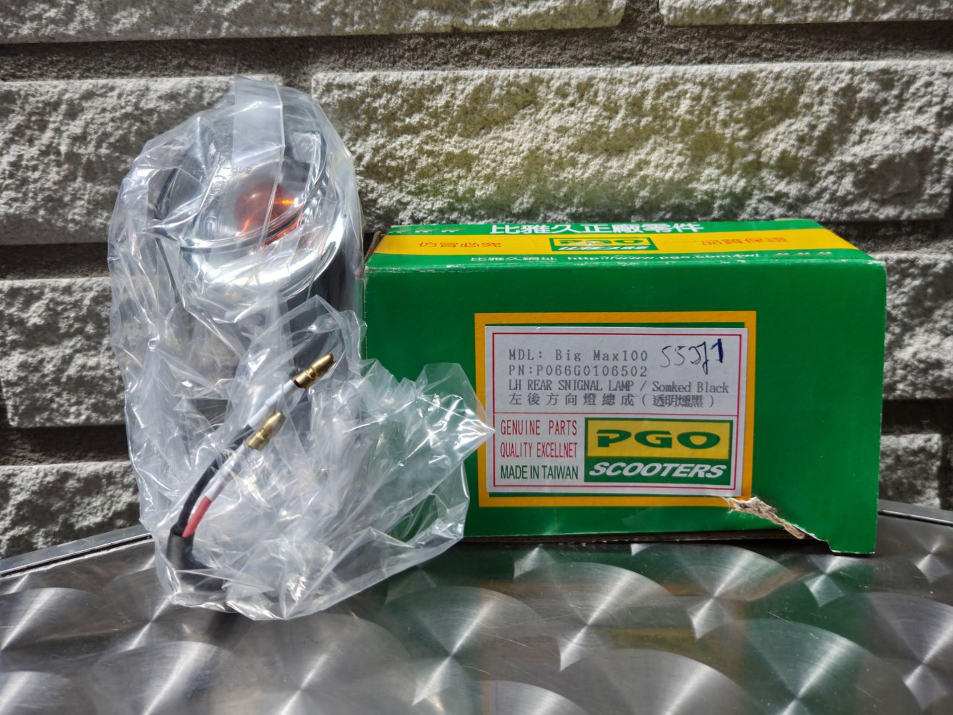 Ingepakt mechanisch onderdeel naast een groene PGO-doos op een metalen oppervlak.