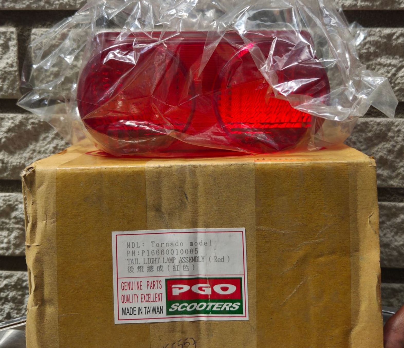 Rood scooterachterlicht, verpakt in plastic, bovenop een kartonnen doos met het opschrift PGO Scooters.