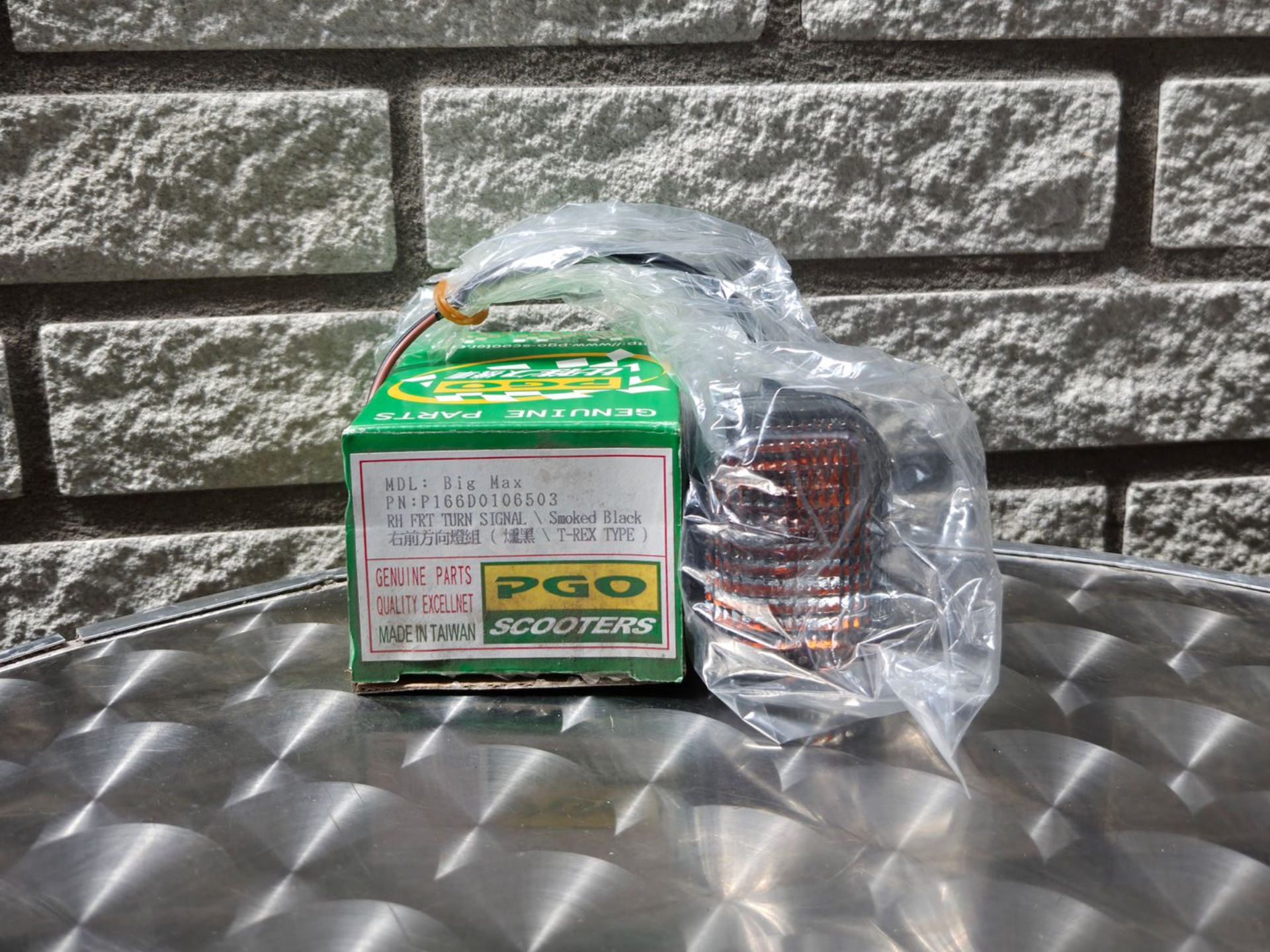 Groene PGO-doos en ingepakt metalen onderdeel op een reflecterende tafel tegen een stenen muur.