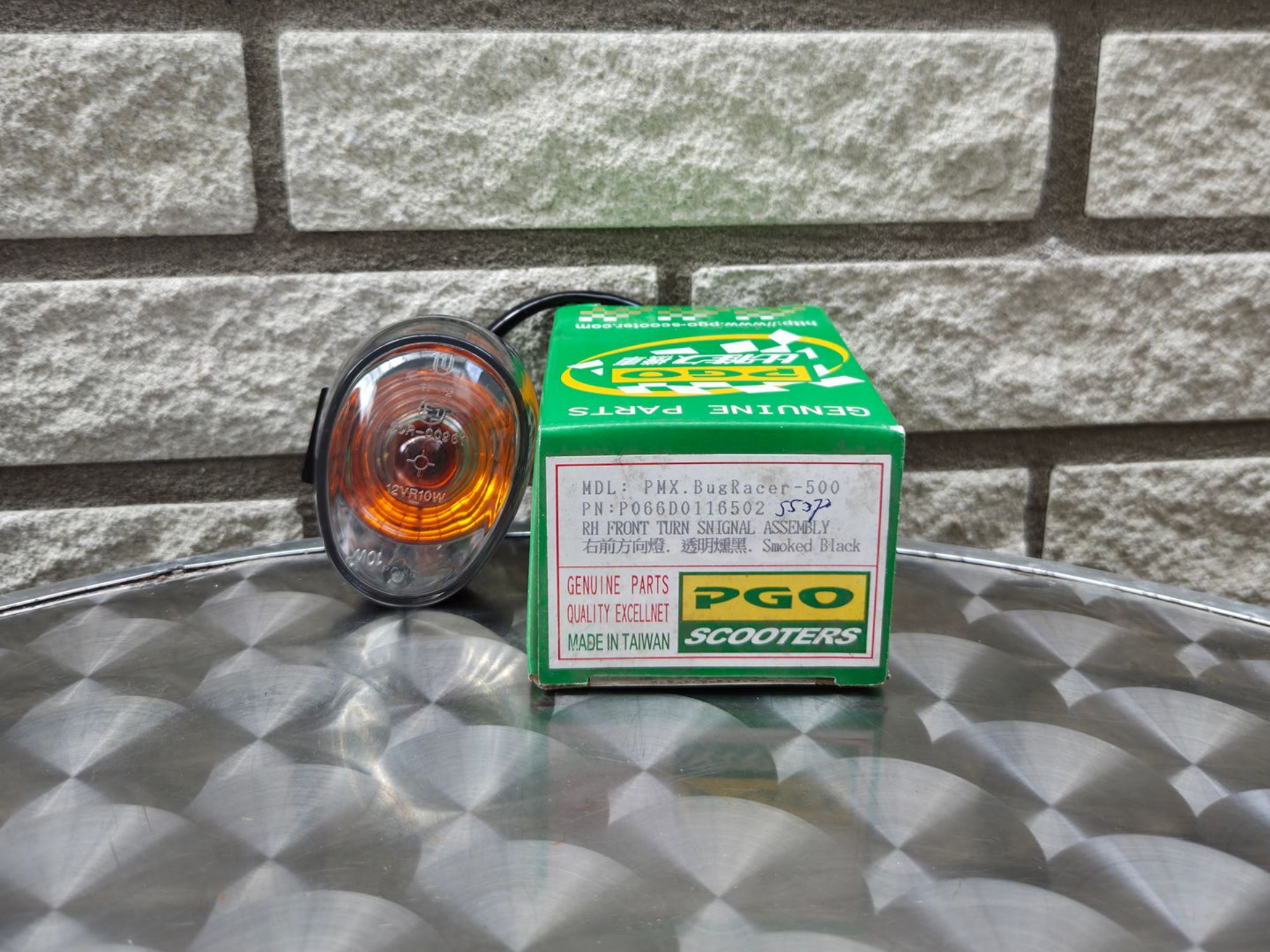 Een oranje autolamp naast een groene PGO-doos op een reflecterend oppervlak tegen een grijze bakstenen muur.