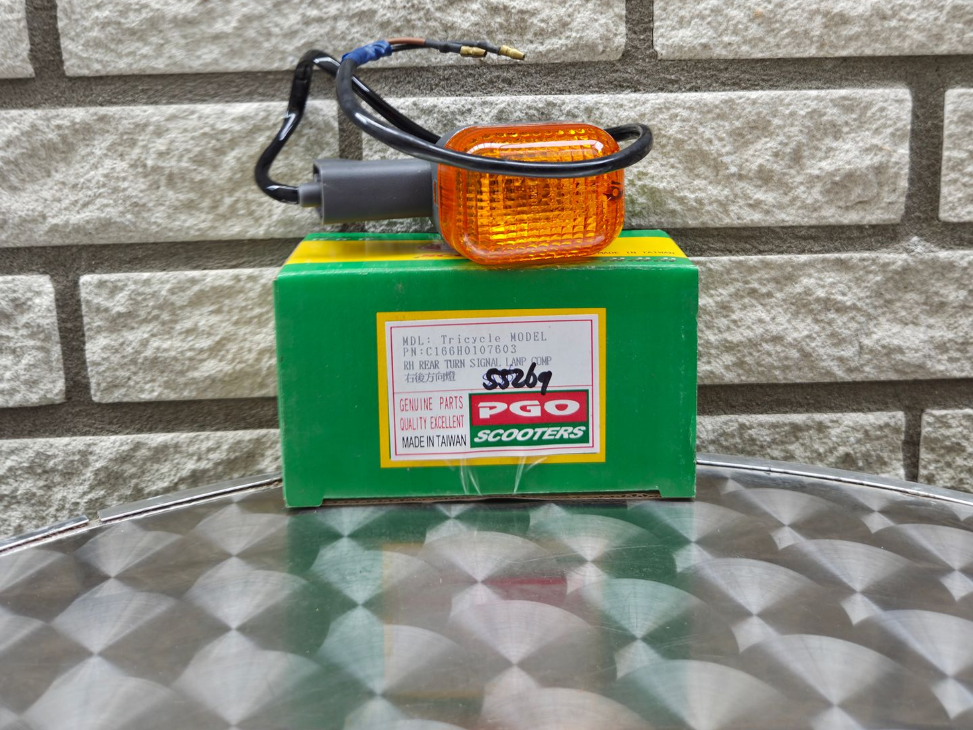 Groene doos met amberkleurig waarschuwingslampje en zwarte kabel op een metalen oppervlak met patroon, tegen een bakstenen muur.