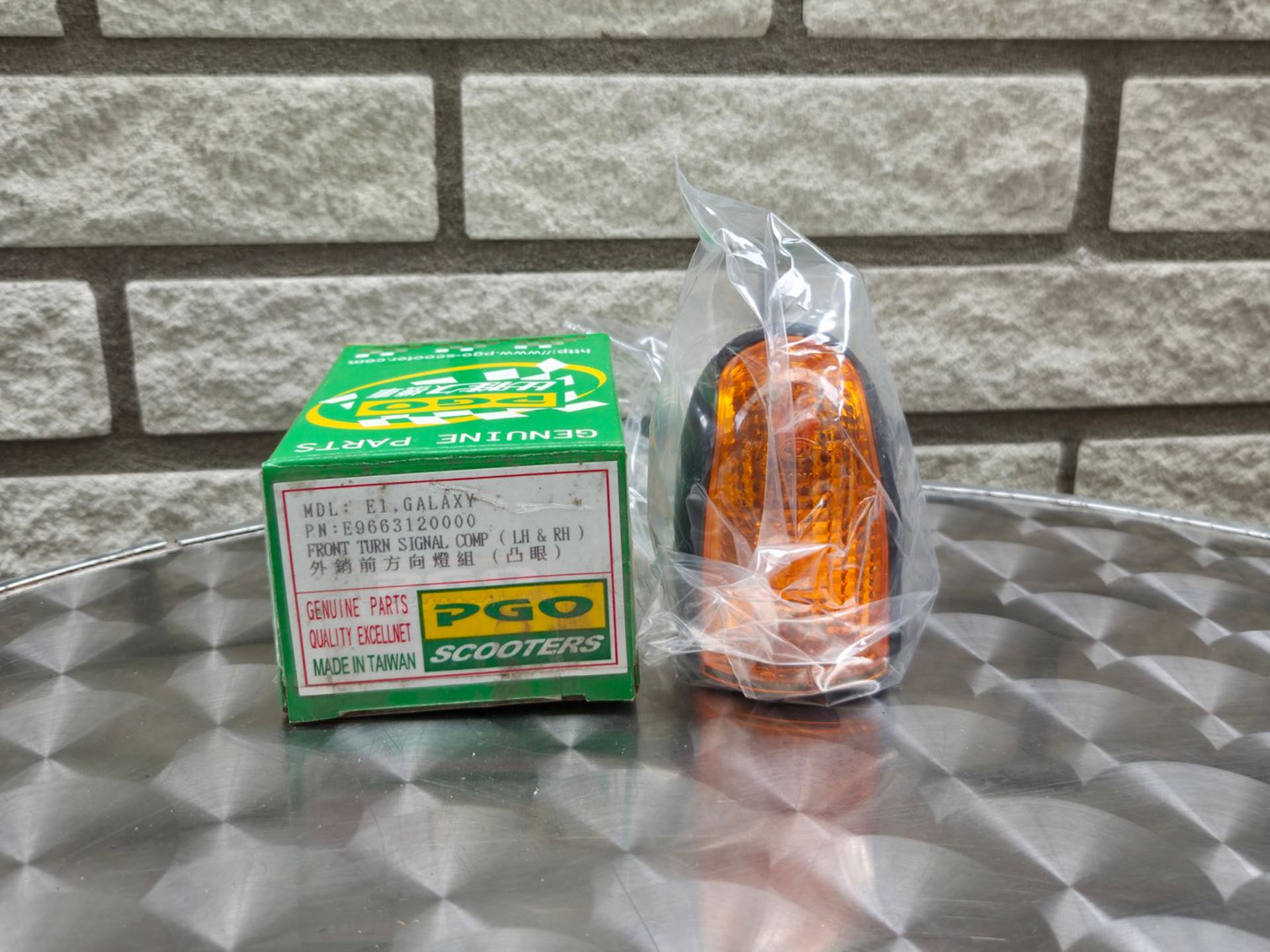 Een groene doos en een oranje speelgoedvoertuig, verpakt in plastic, staan ​​op een metalen tafel tegen een bakstenen muur.