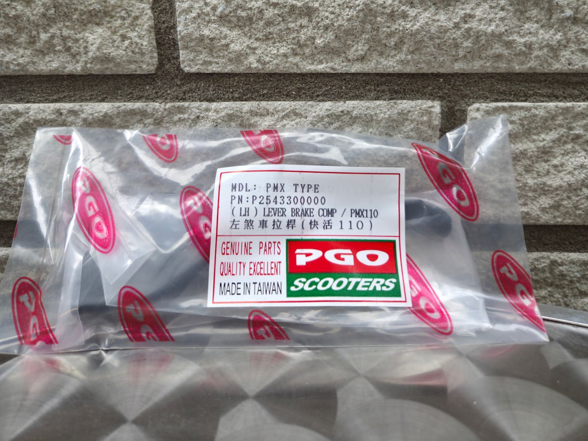 Een in plastic verpakte doos van PGO Scooters tegen een grijze bakstenen muur, met een productetiket erop.