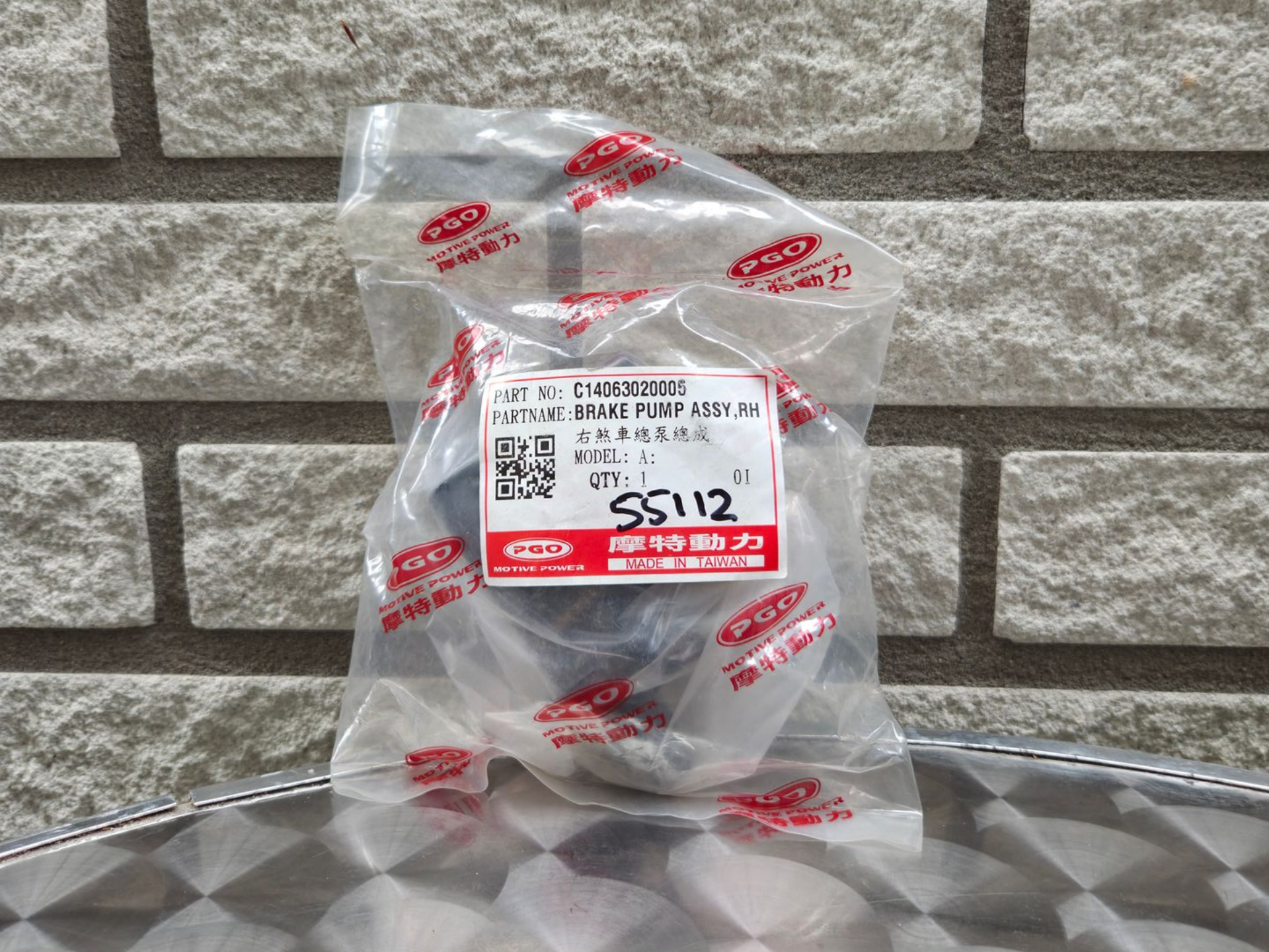 Een doorzichtige plastic winkeltas met een rood-wit etiket ligt op een metalen ondergrond tegen een bakstenen muur.