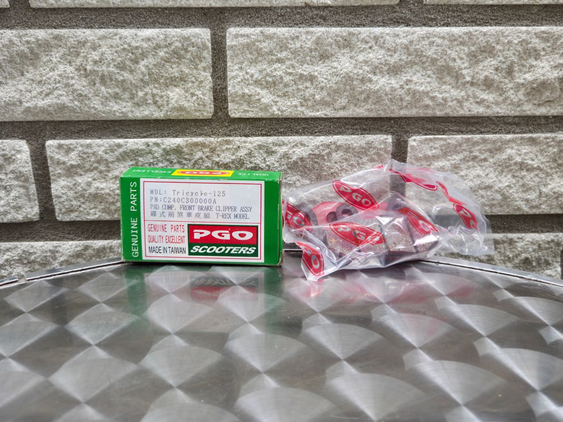 Groene PGO-doos naast een doorzichtige zak met rode capsules op een grijze tegelvloer tegen een bakstenen muur.