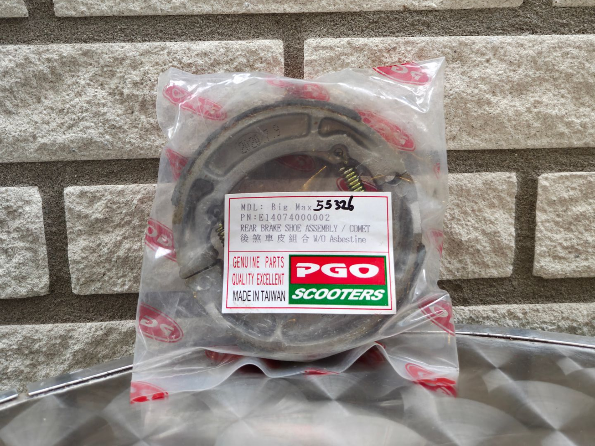 Een rond PGO-scooteronderdeel, verpakt in transparant plastic, ligt op een grijze ondergrond tegen een bakstenen muur.