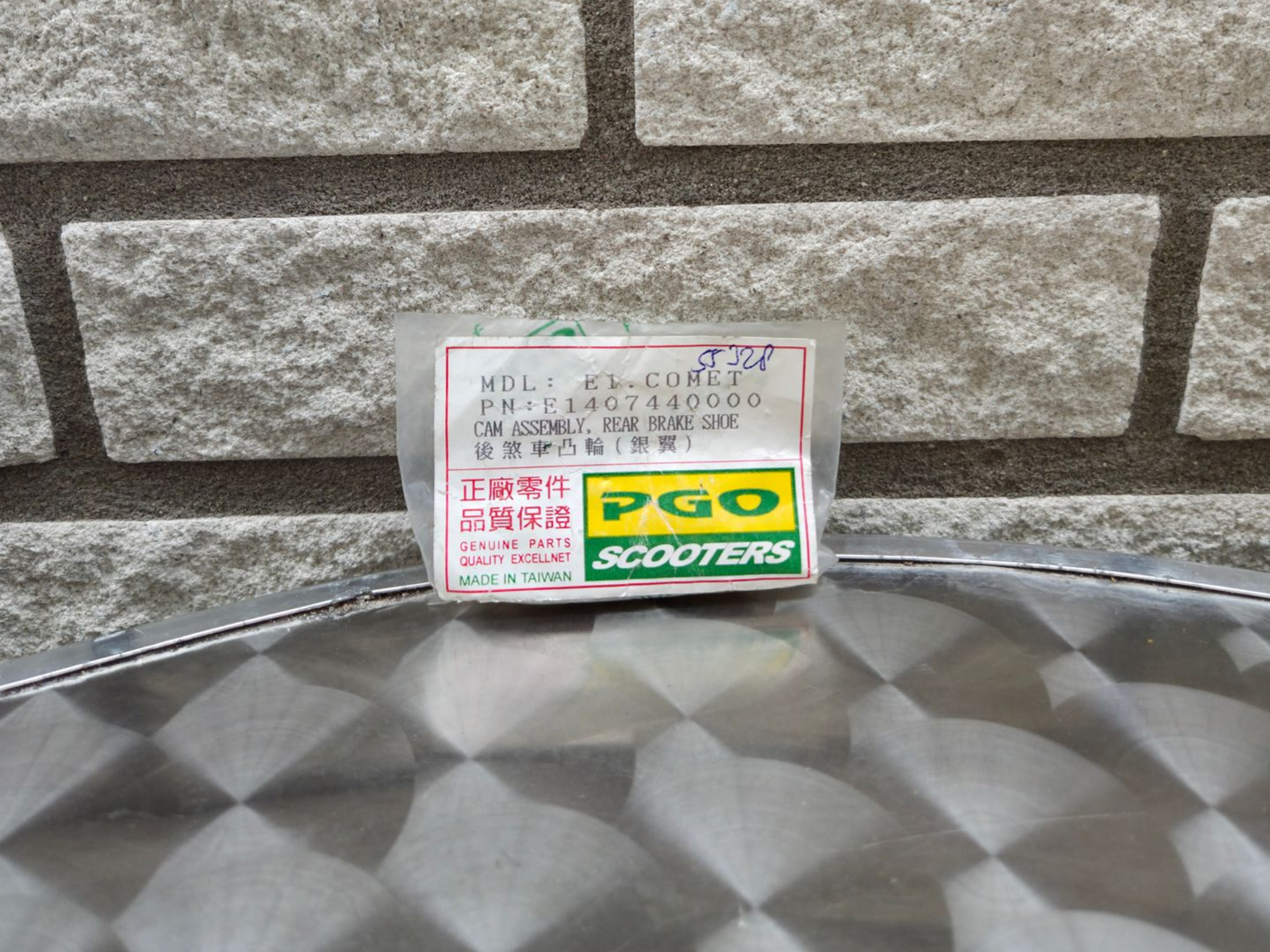 Klein PGO Scooters-logo op een grijze stenen muur boven een metalen oppervlak met patroon.