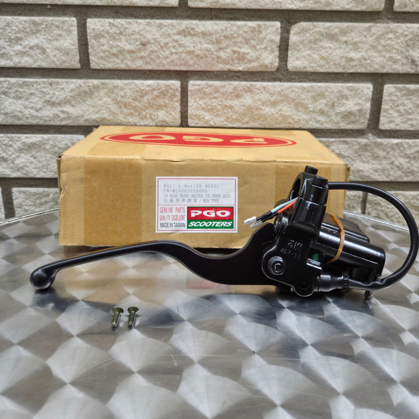Zwarte motorfietsremhendel en hoofdremcilinder op een metalen ondergrond, met daarachter een kartonnen doos.