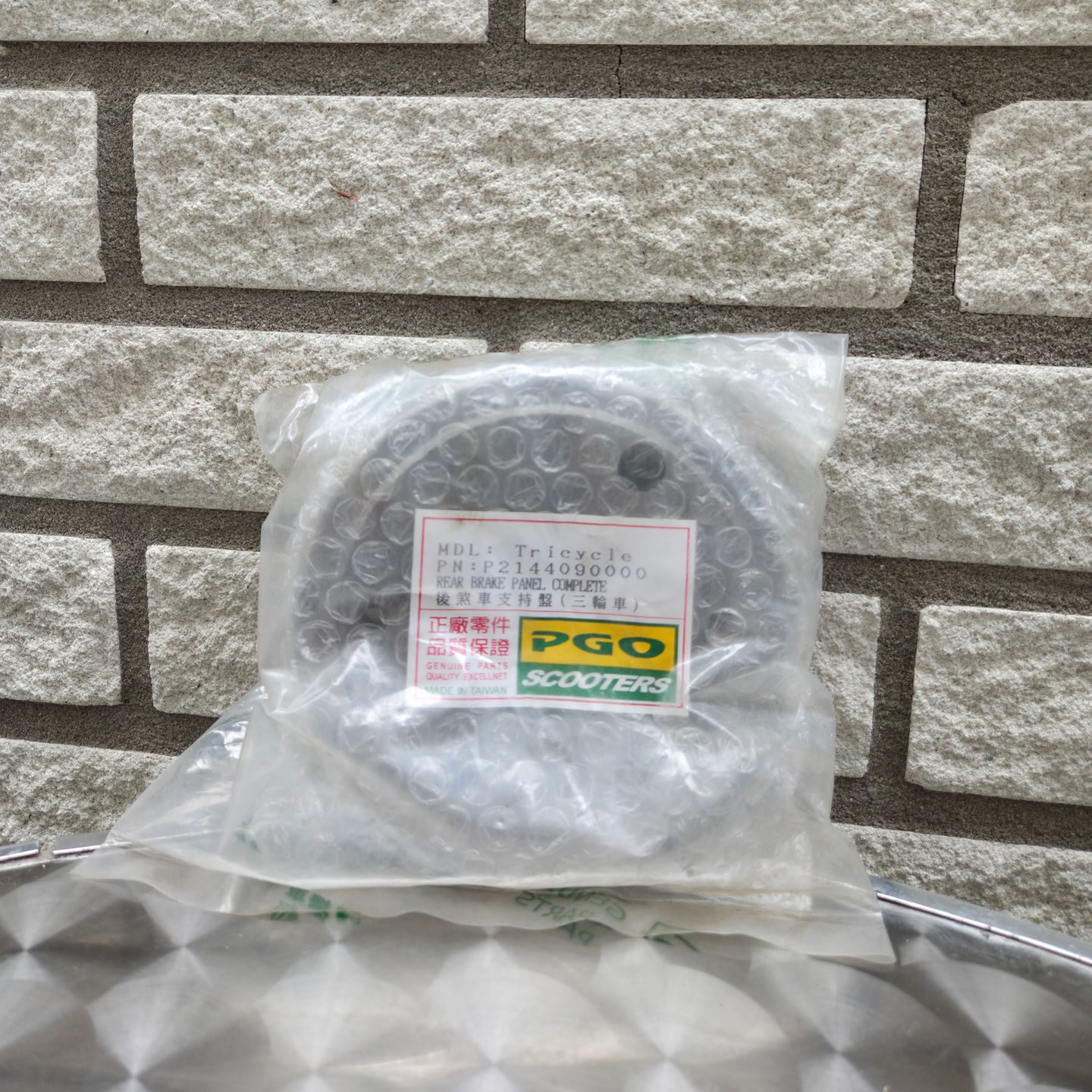 Een in plastic verpakt, rond, donkermetalen onderdeel met een geel etiket, dat tegen een bakstenen muur leunt.