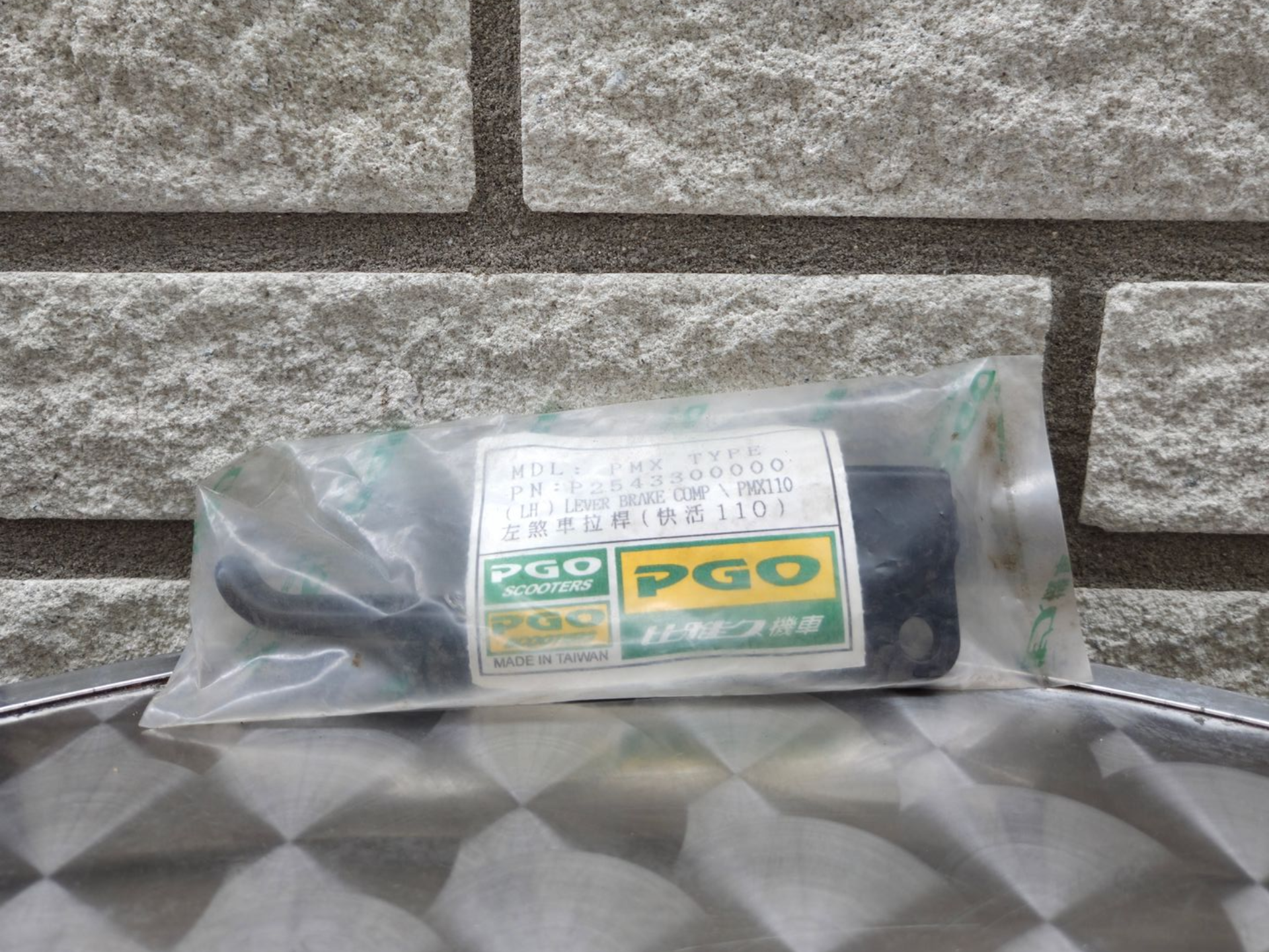 Een klein, doorzichtig plastic zakje met een groen PGO-etiket, dat op een zilverkleurige, gestructureerde ondergrond tegen een bakstenen muur rust.