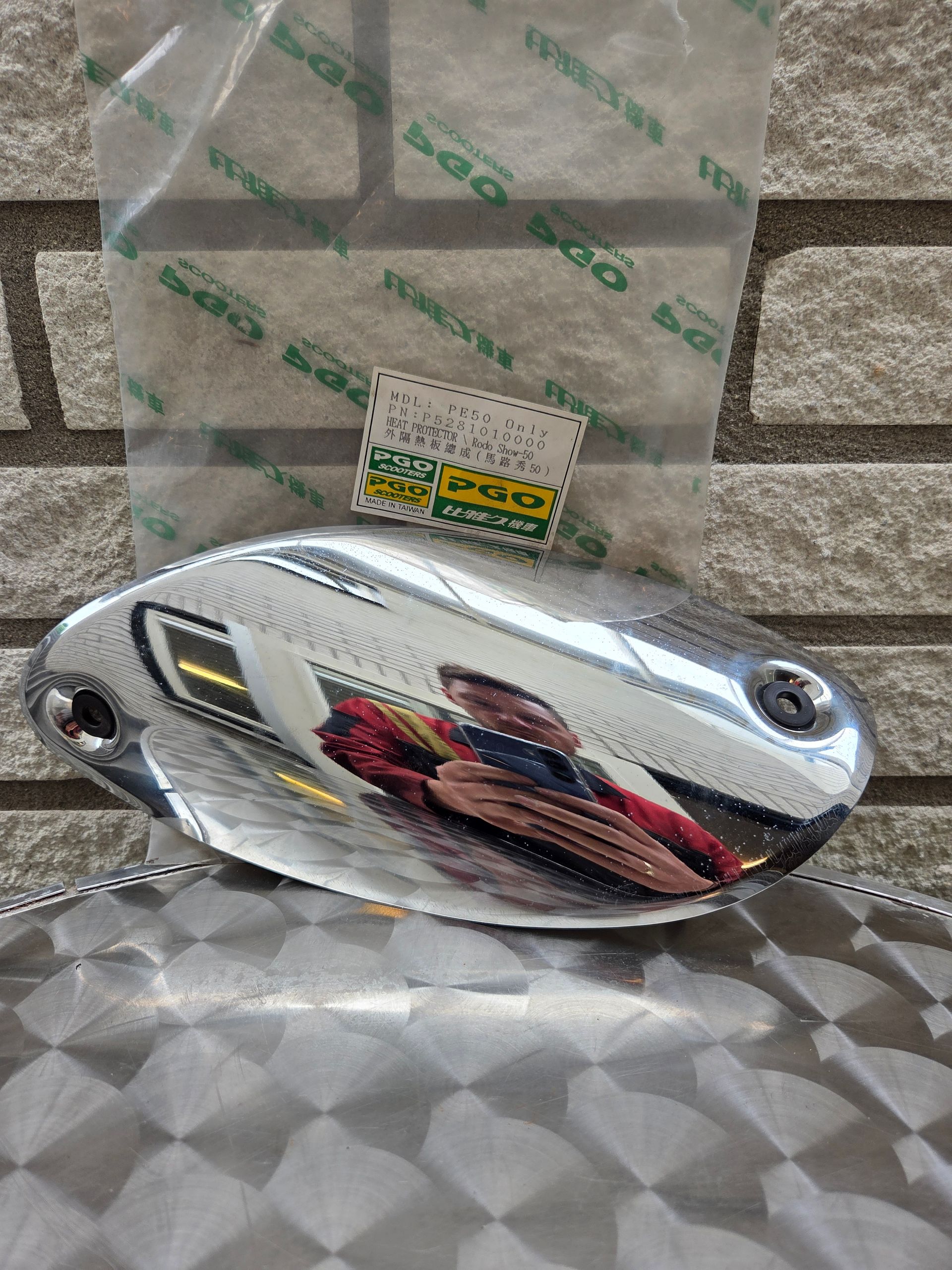 Een ovaal verchroomd motoronderdeel met een reflecterend oppervlak, liggend op een metalen tafel met patroon en een prijskaartje erachter.