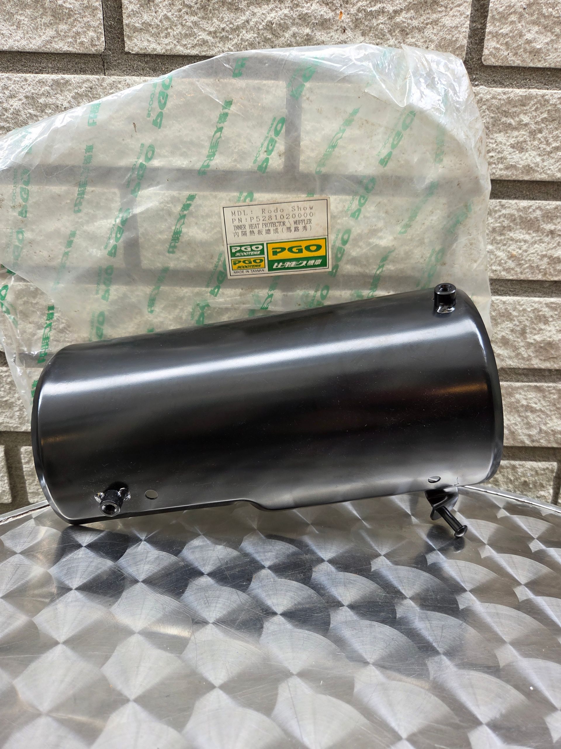 Een zwart cilindrisch metalen onderdeel rust op een vloer met patroon voor een ingepakt pakket.
