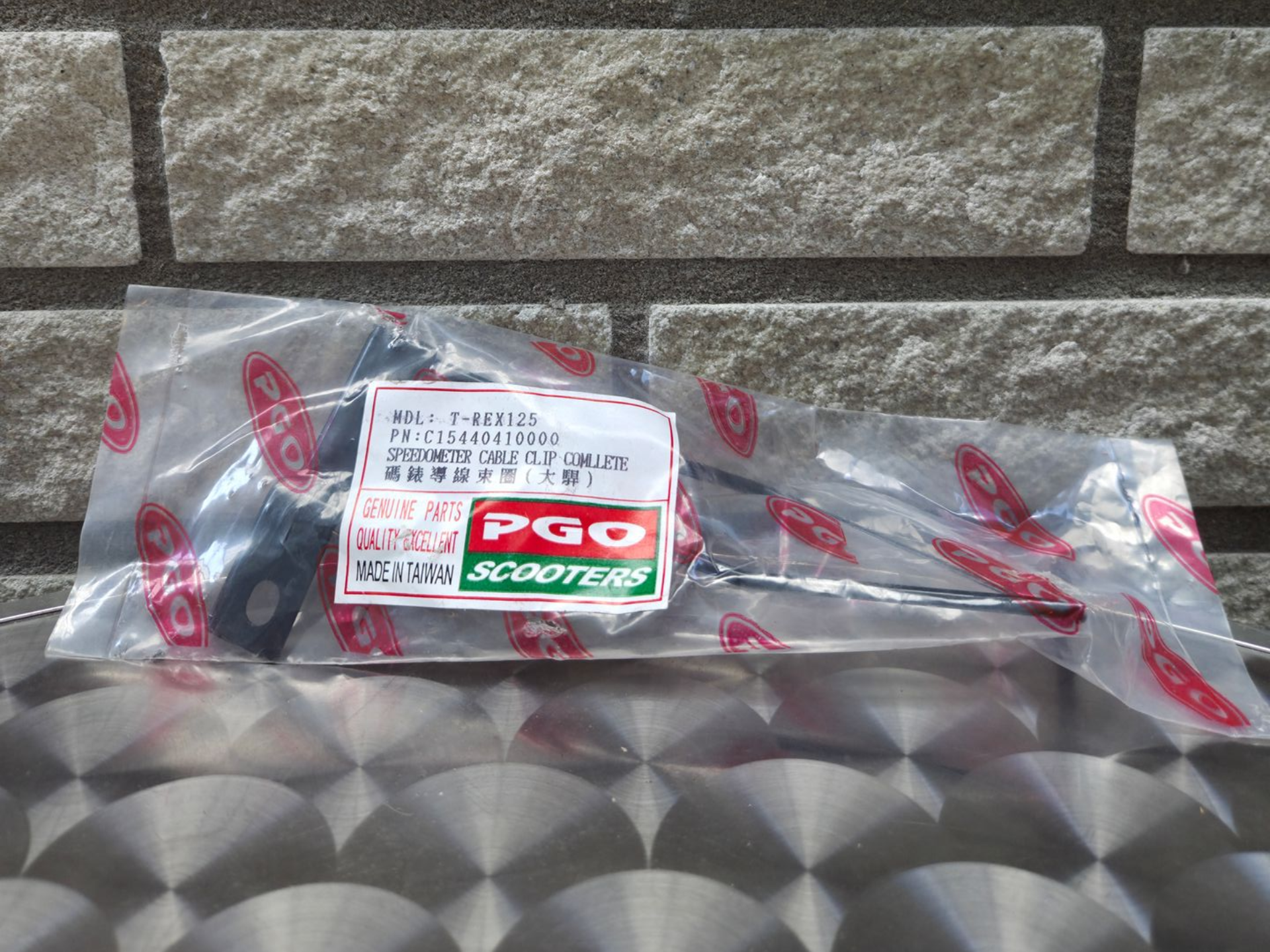 In plastic verpakt PGO-motoronderdeel op een oppervlak met patroon tegen een stenen muur.