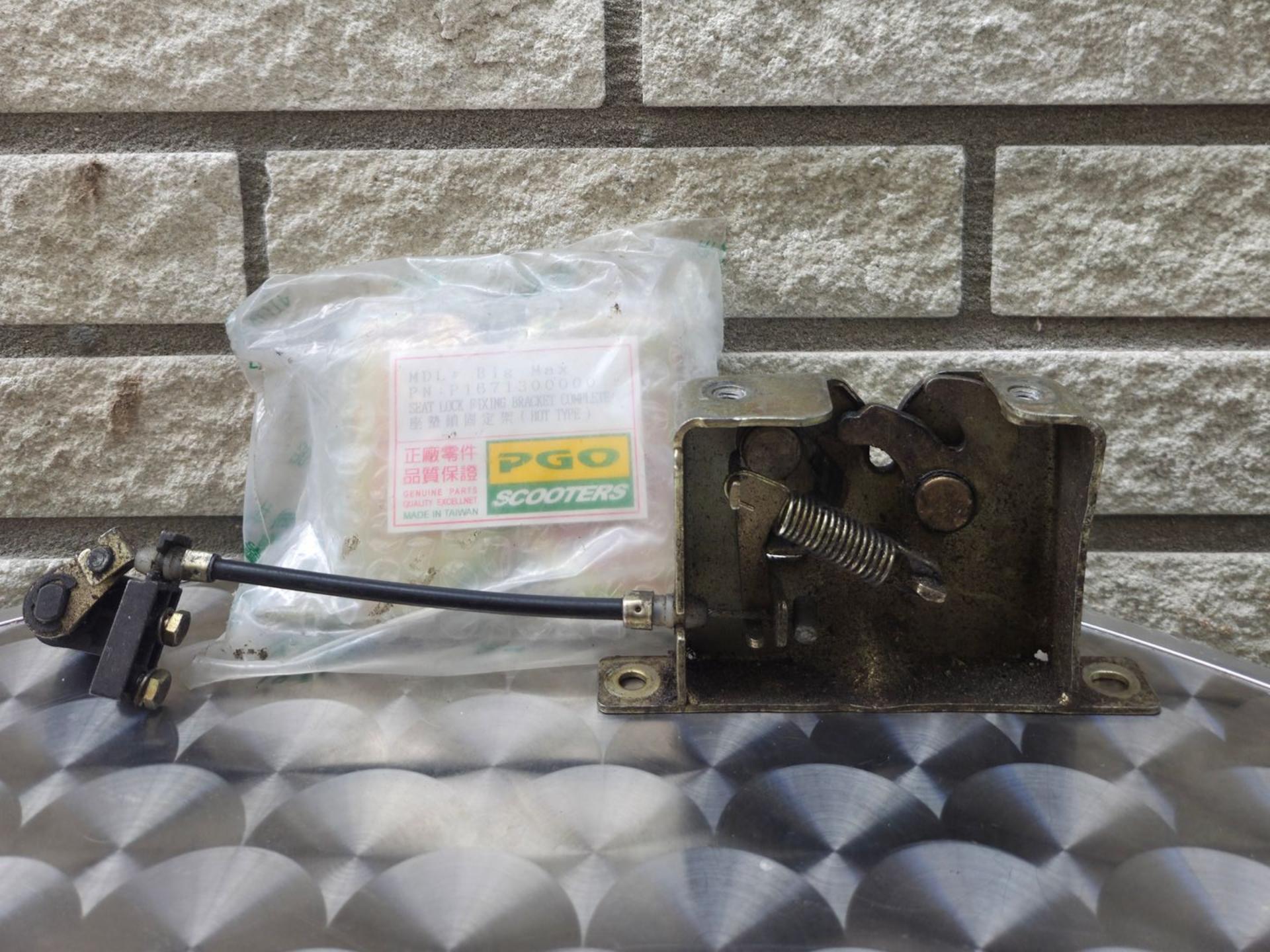Metalen sluitmechanisme met aangehechte stang naast een klein plastic zakje, op een oppervlak met patroon tegen een bakstenen muur.