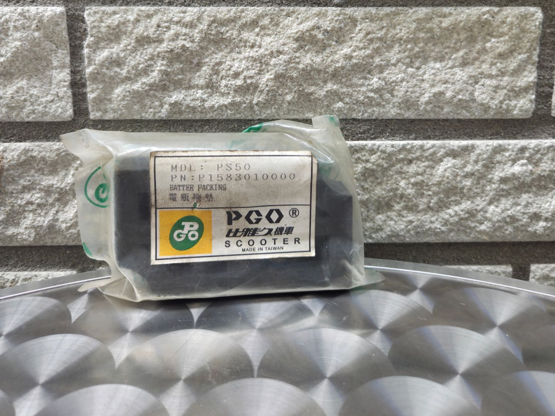 Ingepakt industrieel onderdeel met een GE/PCO-label, rustend op een zilverkleurig, gestructureerd oppervlak tegen een bakstenen muur.
