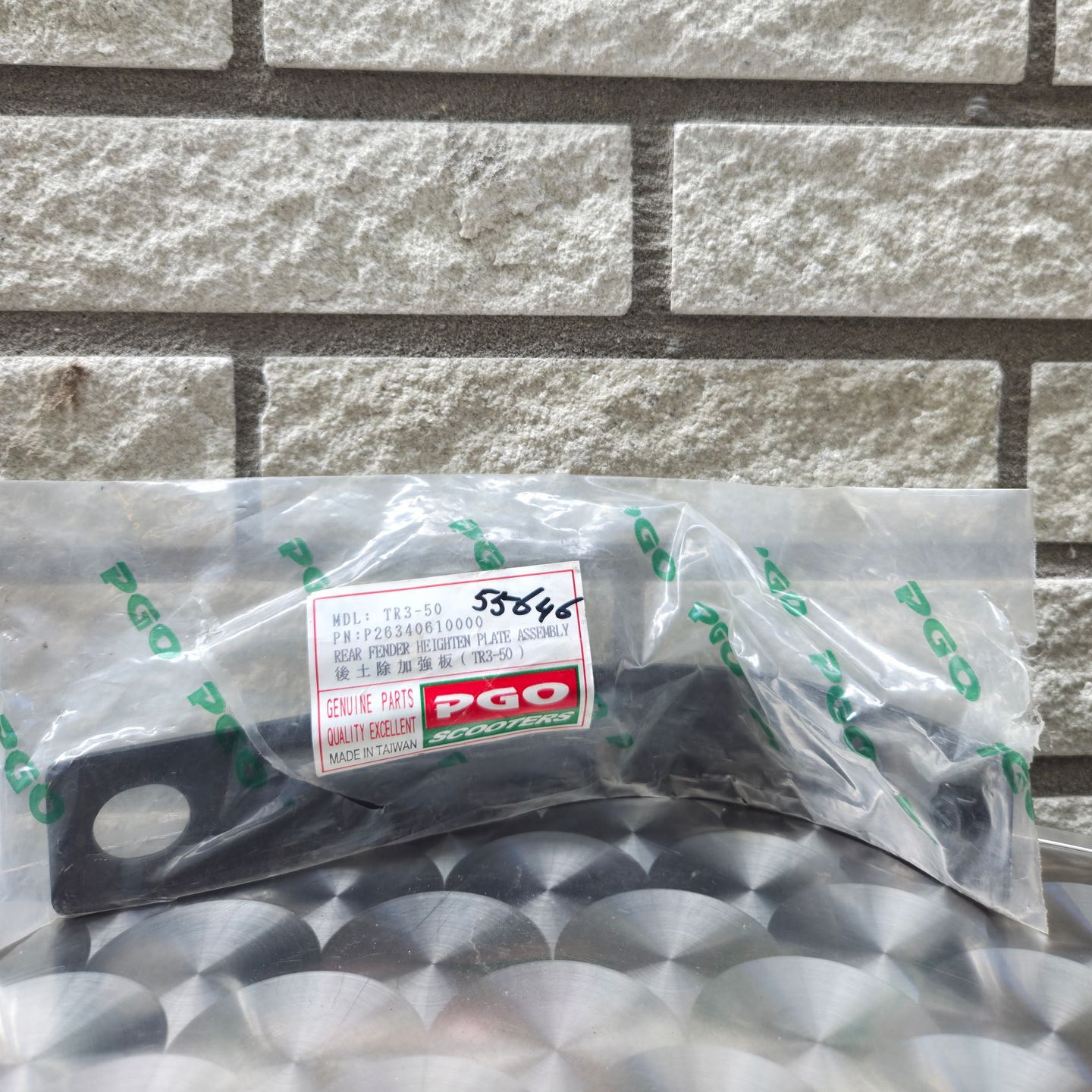 Een plastic auto-onderdeel in een transparante verpakking op een zilverkleurige ondergrond tegen een bakstenen muur.